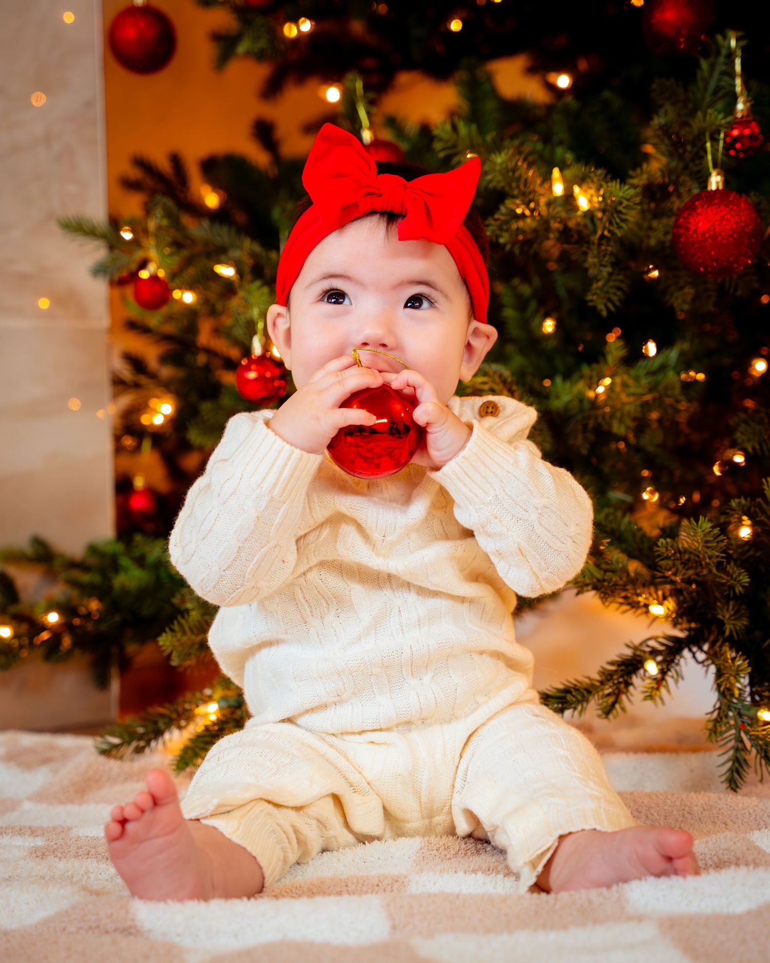 Baby in cream-colored dress sits by Christmas tree with red bow and ornaments in festive holiday scene.
