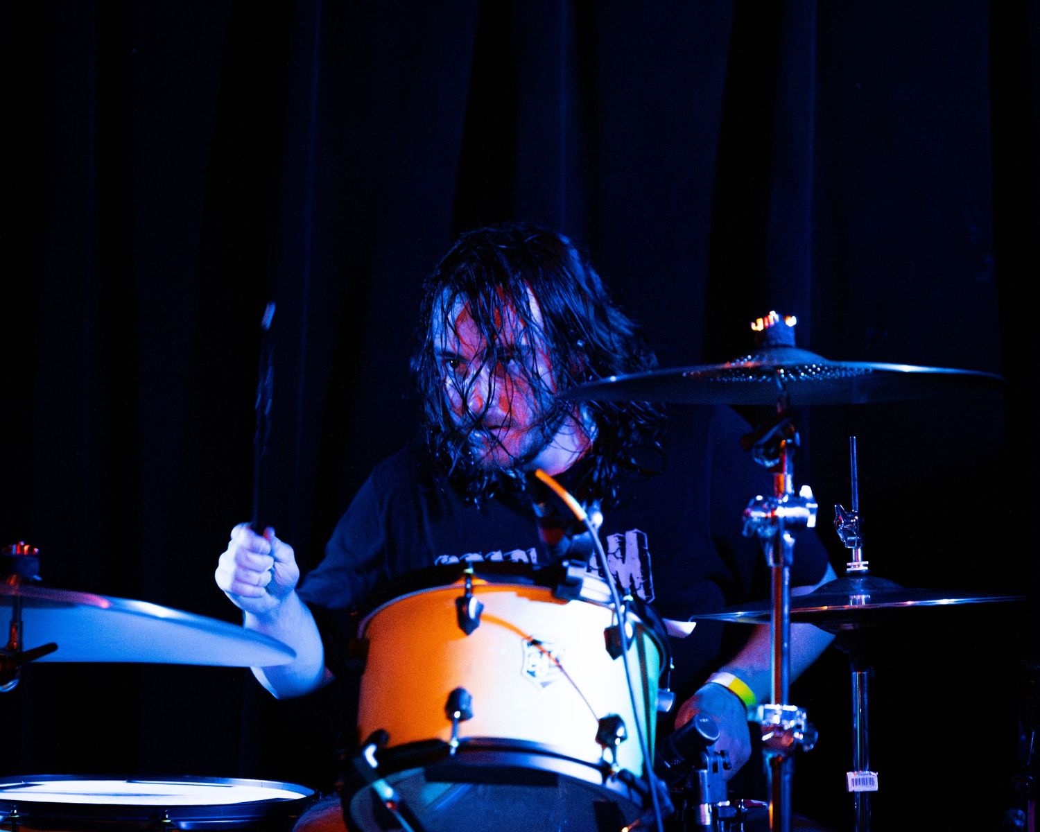 Drummer plays orange drum kit on dark stage with dramatic backlighting.