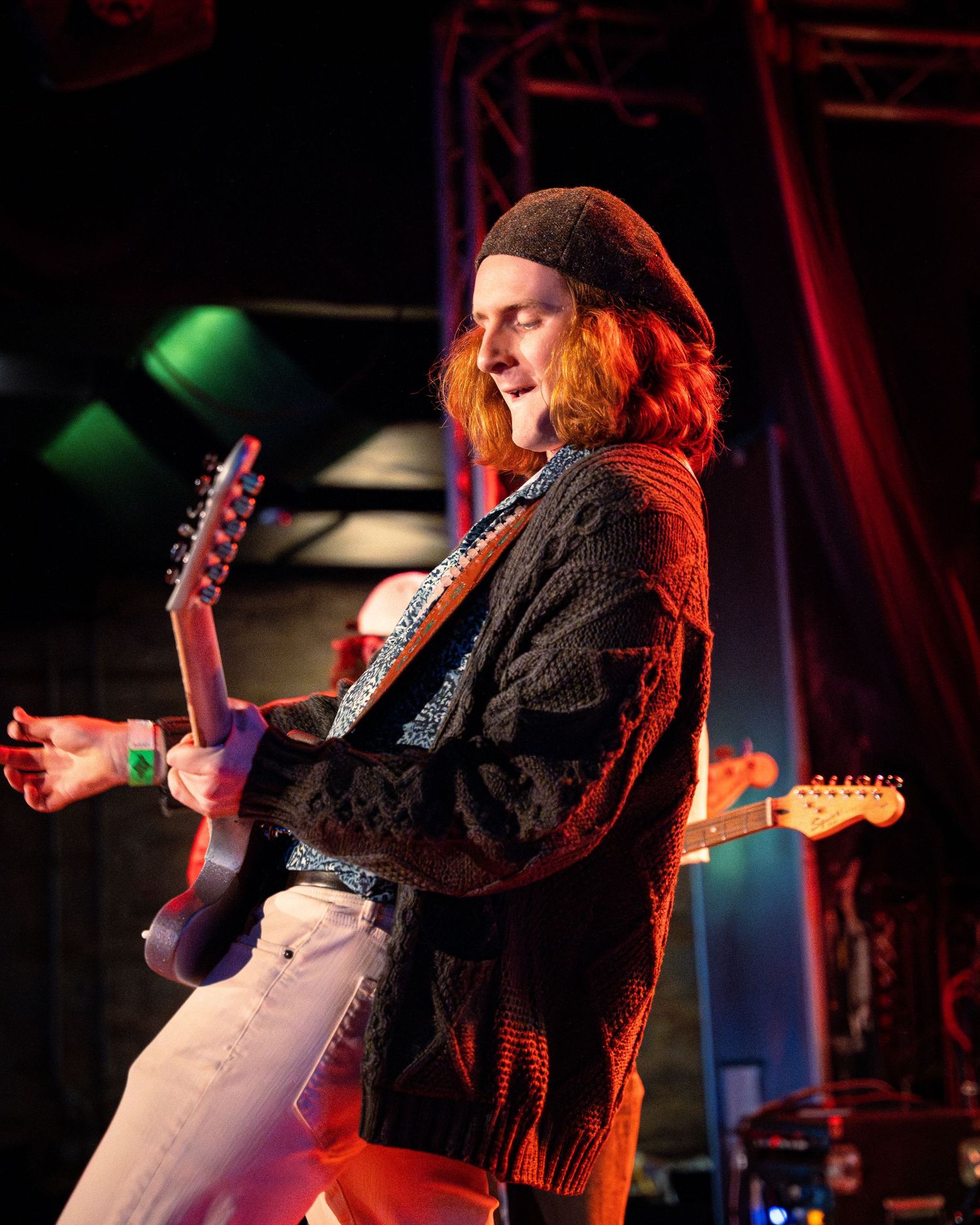 Musician plays electric guitar on stage under dramatic red and green lighting during live performance.
