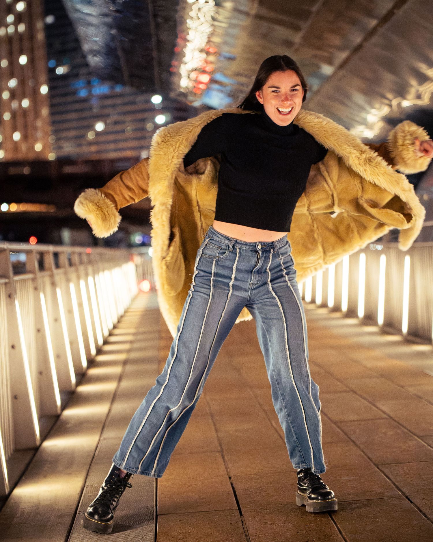 Person wearing a furry coat and striped pants dances playfully in a tunnel lit by warm lights.