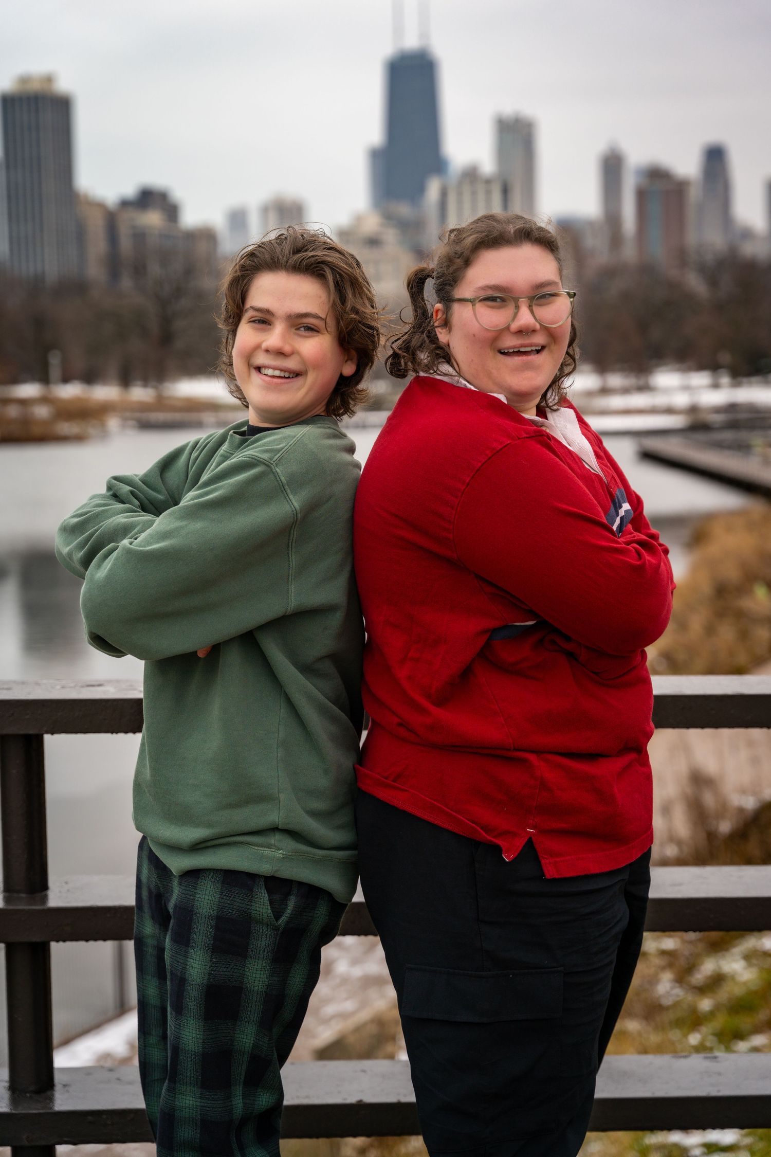 Two friends in winter clothing pose together on a lakefront with the Chicago skyline visible in the background.