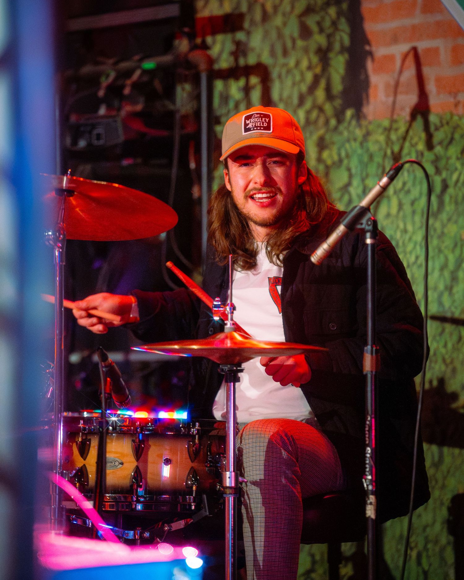Drummer in orange cap performing behind drum kit with colored stage lights.