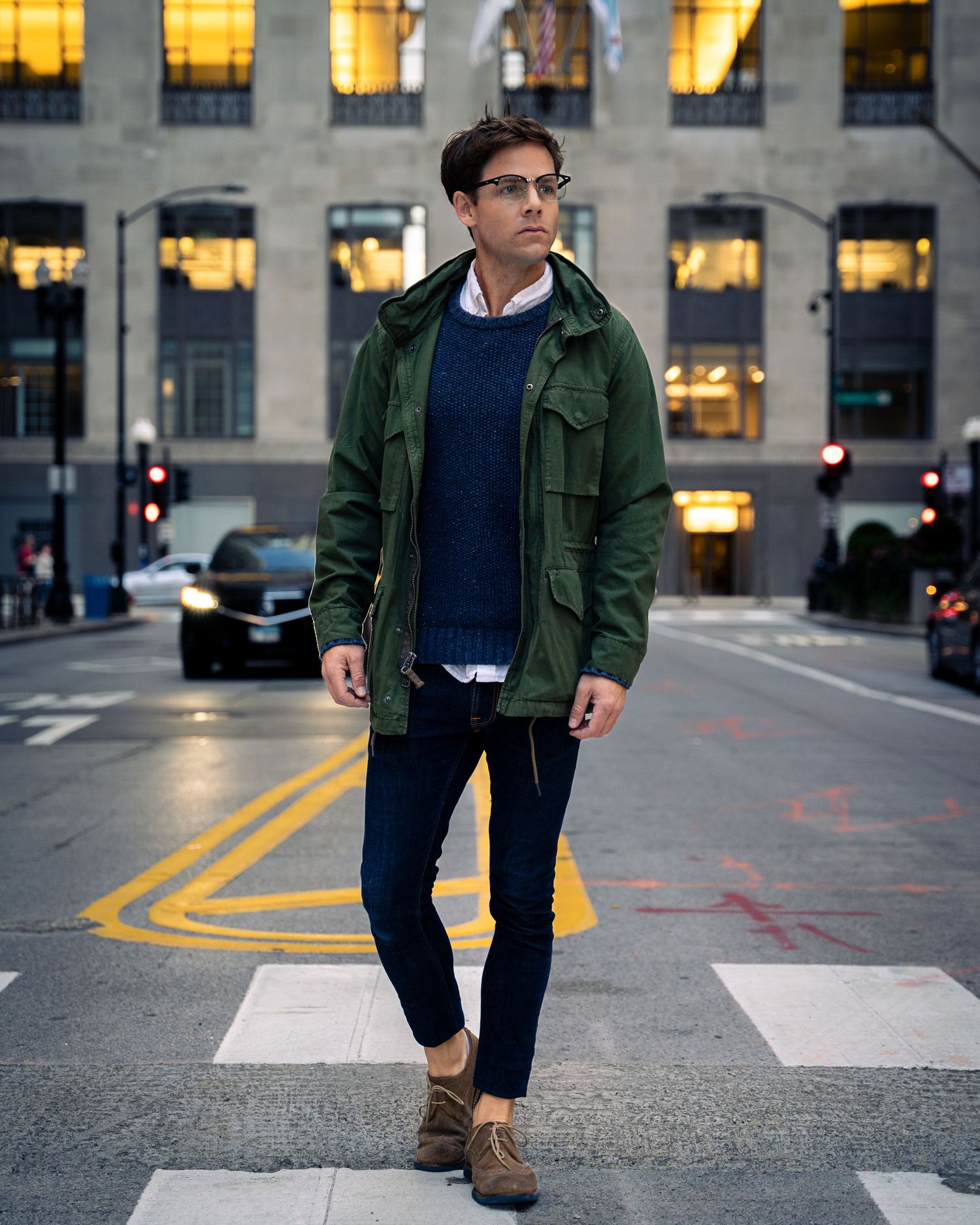 Street style photography at dusk featuring olive jacket and dark denim against city backdrop.
