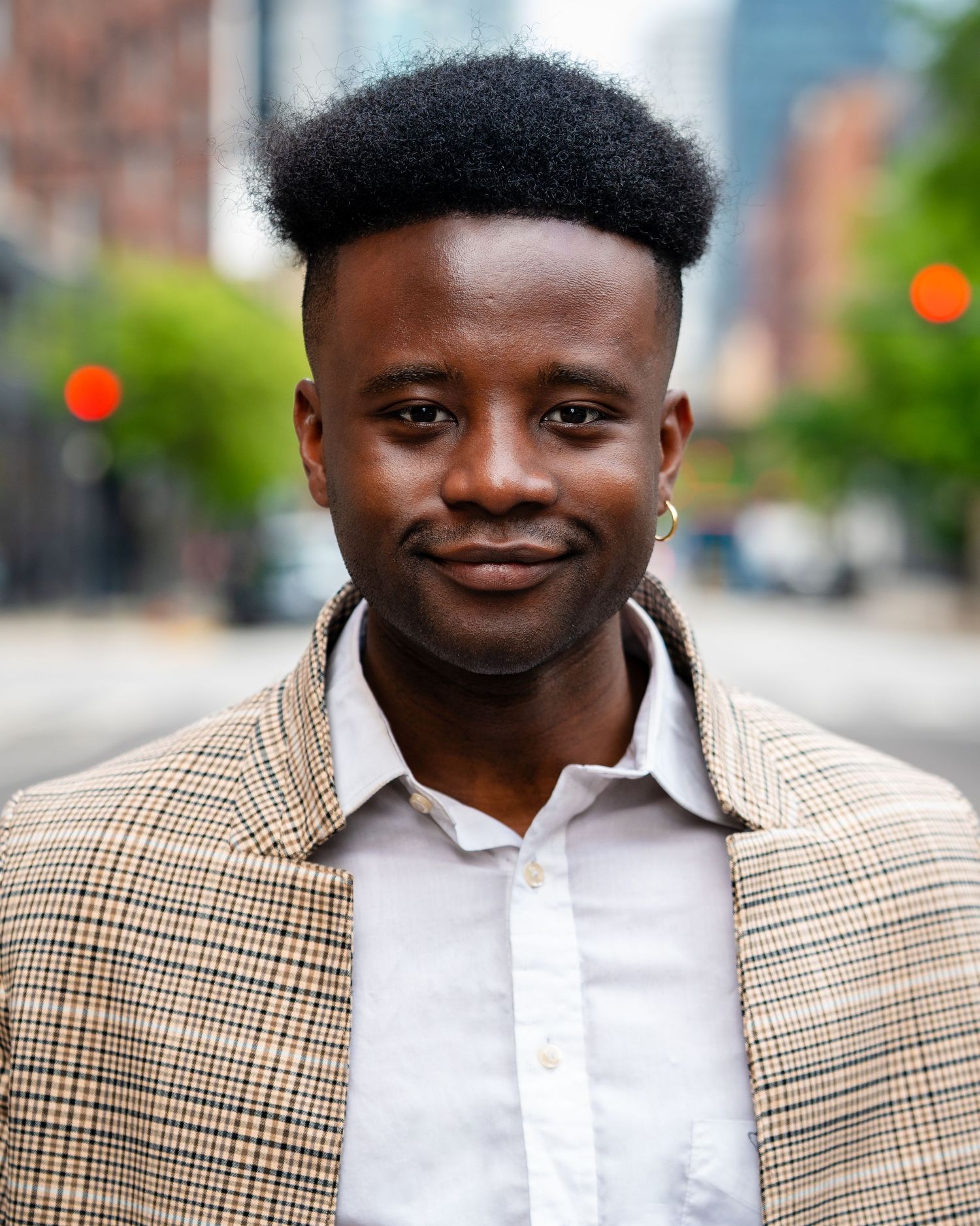 Professional portrait of a person in a plaid jacket and white shirt smiling outdoors in an urban setting.