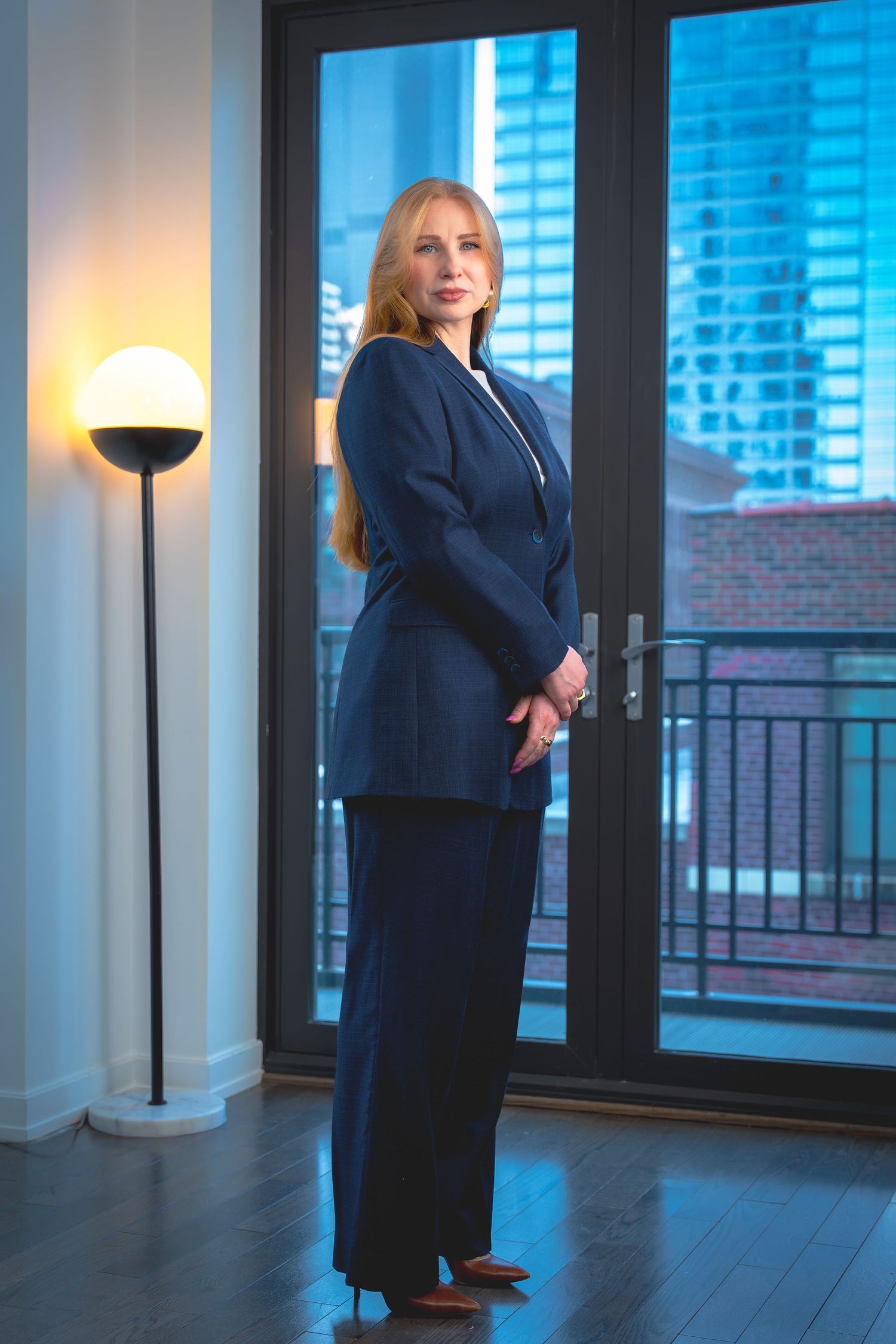 Business professional in a navy suit stands near large windows in modern office space at dusk with city views.