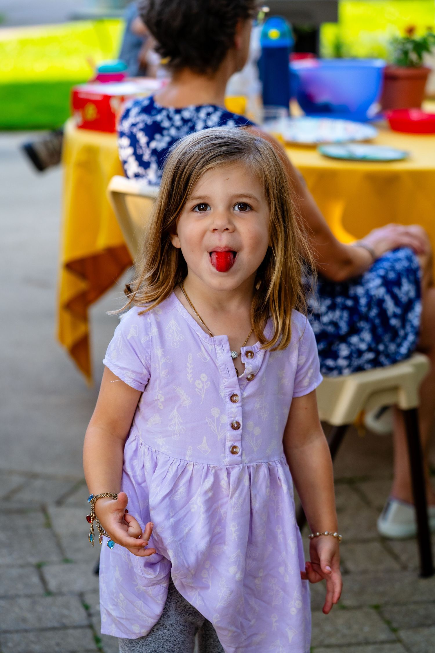 A child in a purple dress makes silly expressions while playing outdoors at a colorful party setting.