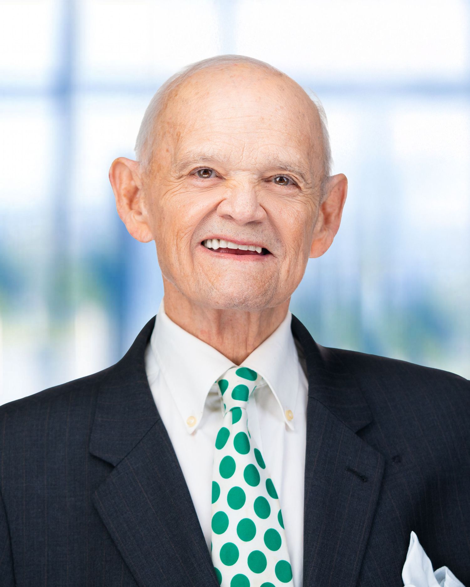 Professional headshot of senior executive wearing black suit and green polka dot tie against bright background.