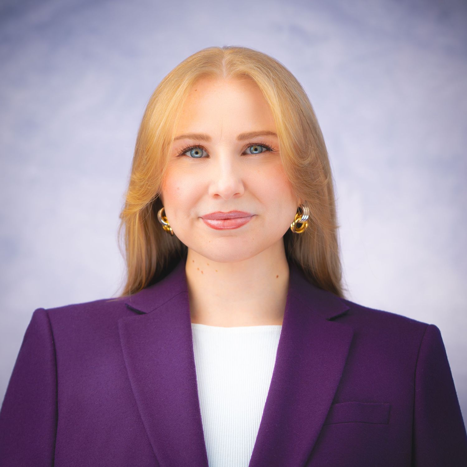 Professional headshot of a person wearing a purple blazer and white top against a light gray background.