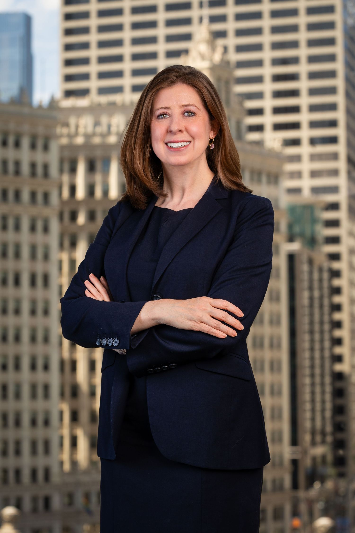 Professional poses with crossed arms in dark blazer against city skyline.