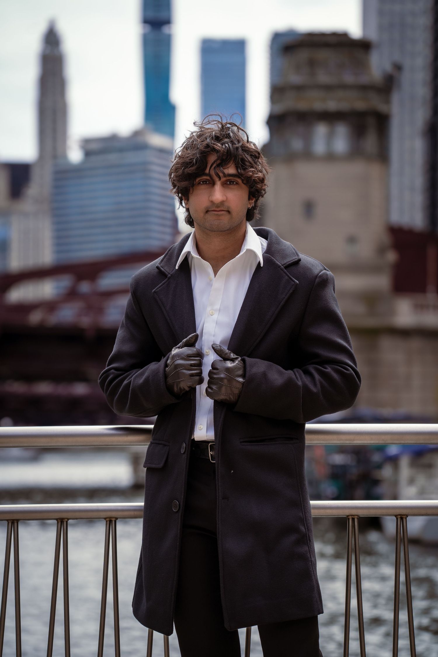 Person in formal black suit and white shirt standing by waterfront with city skyline.
