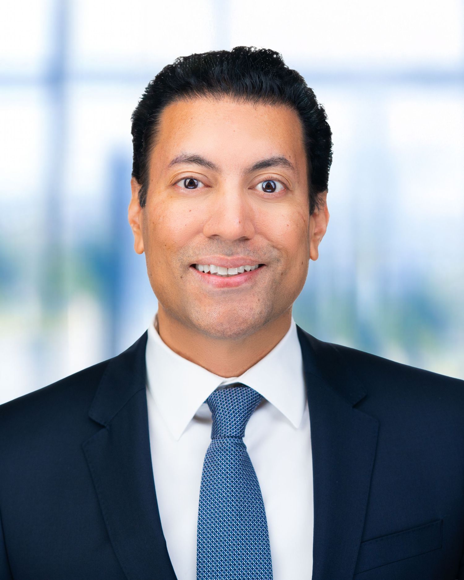 Corporate portrait of person in dark suit and blue patterned tie against modern office setting.