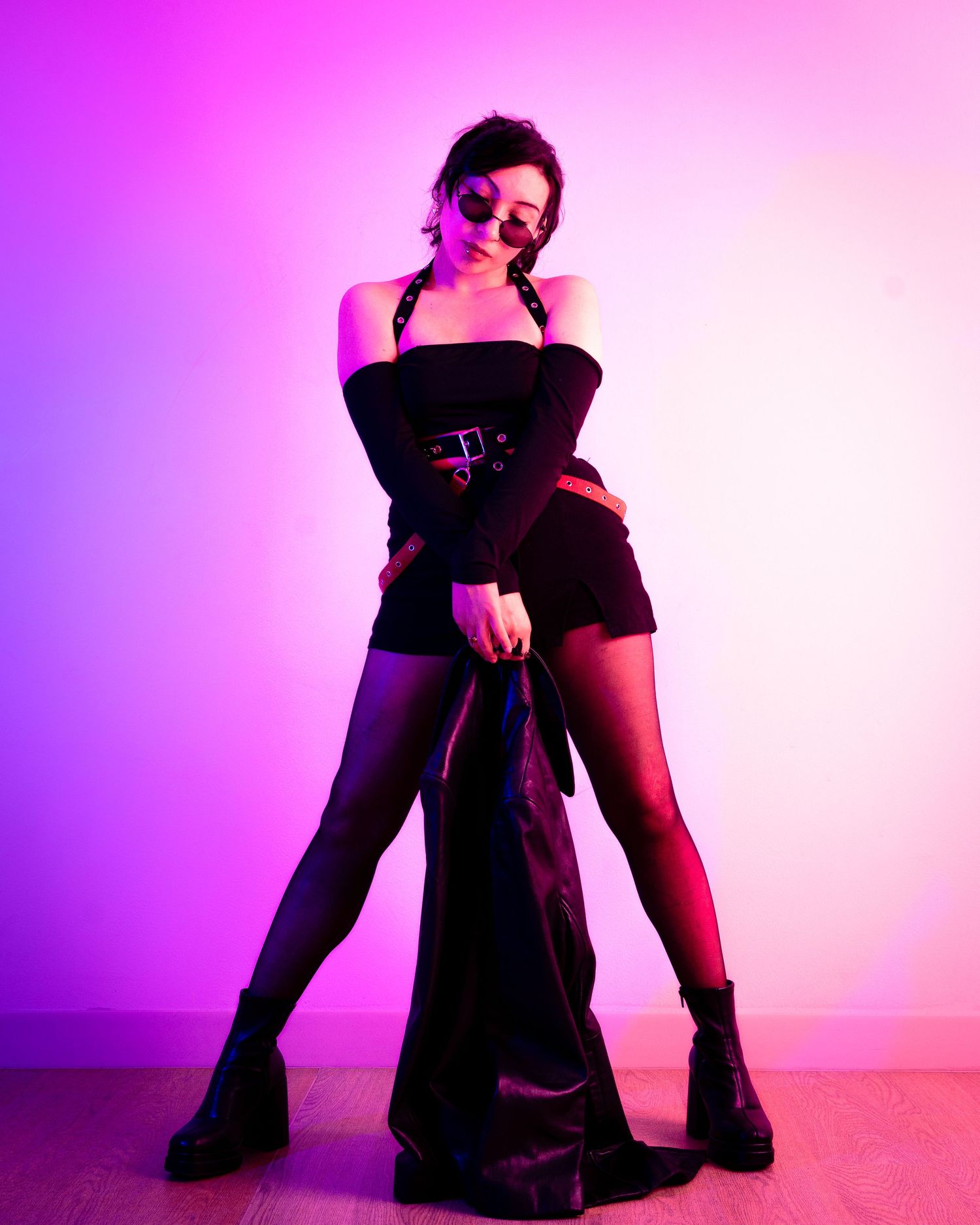 Dramatic fashion pose in black outfit and boots against gradient purple-pink backdrop.