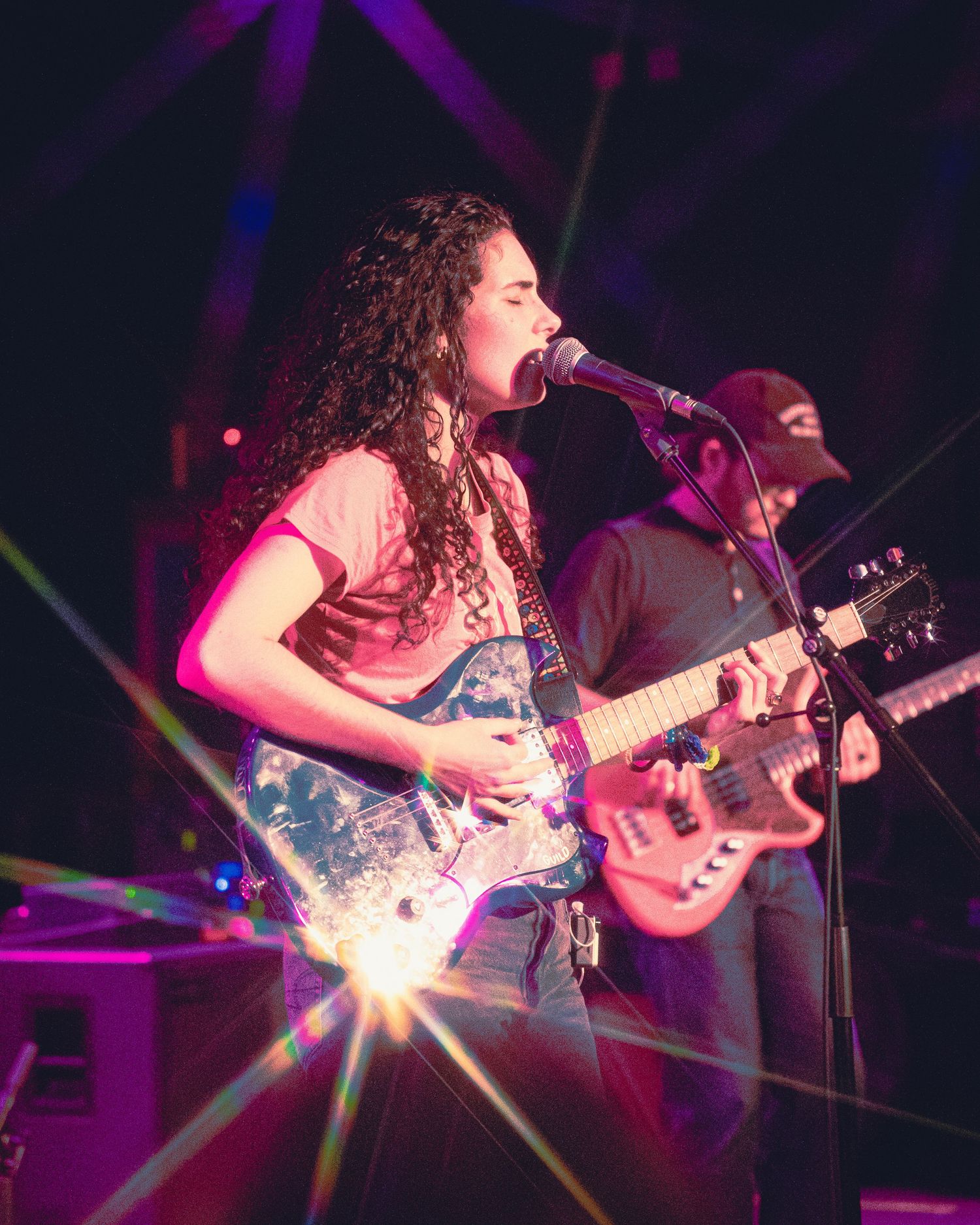A musician performs on stage with curly hair and a guitar under colorful purple and blue stage lighting.