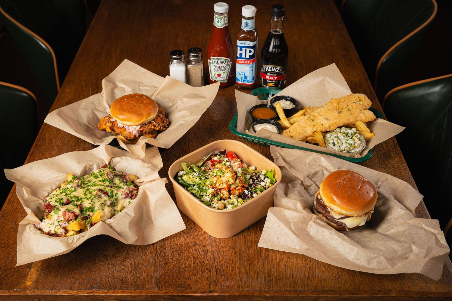 Array of gourmet burgers and salads served on kraft paper at a rustic wooden table.