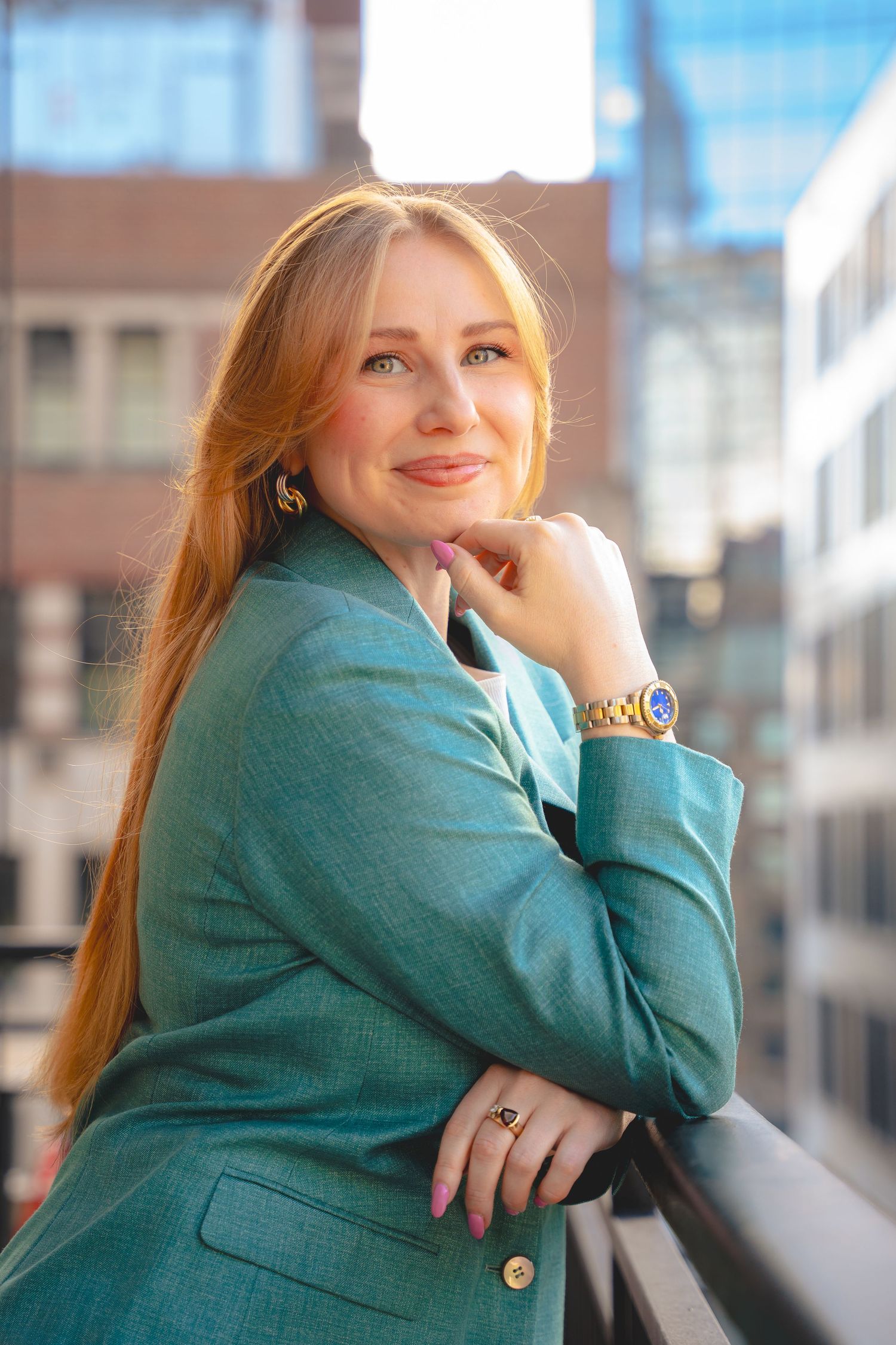 Portrait of a woman in a turquoise blazer leaning on a railing with sunlit buildings in the background.