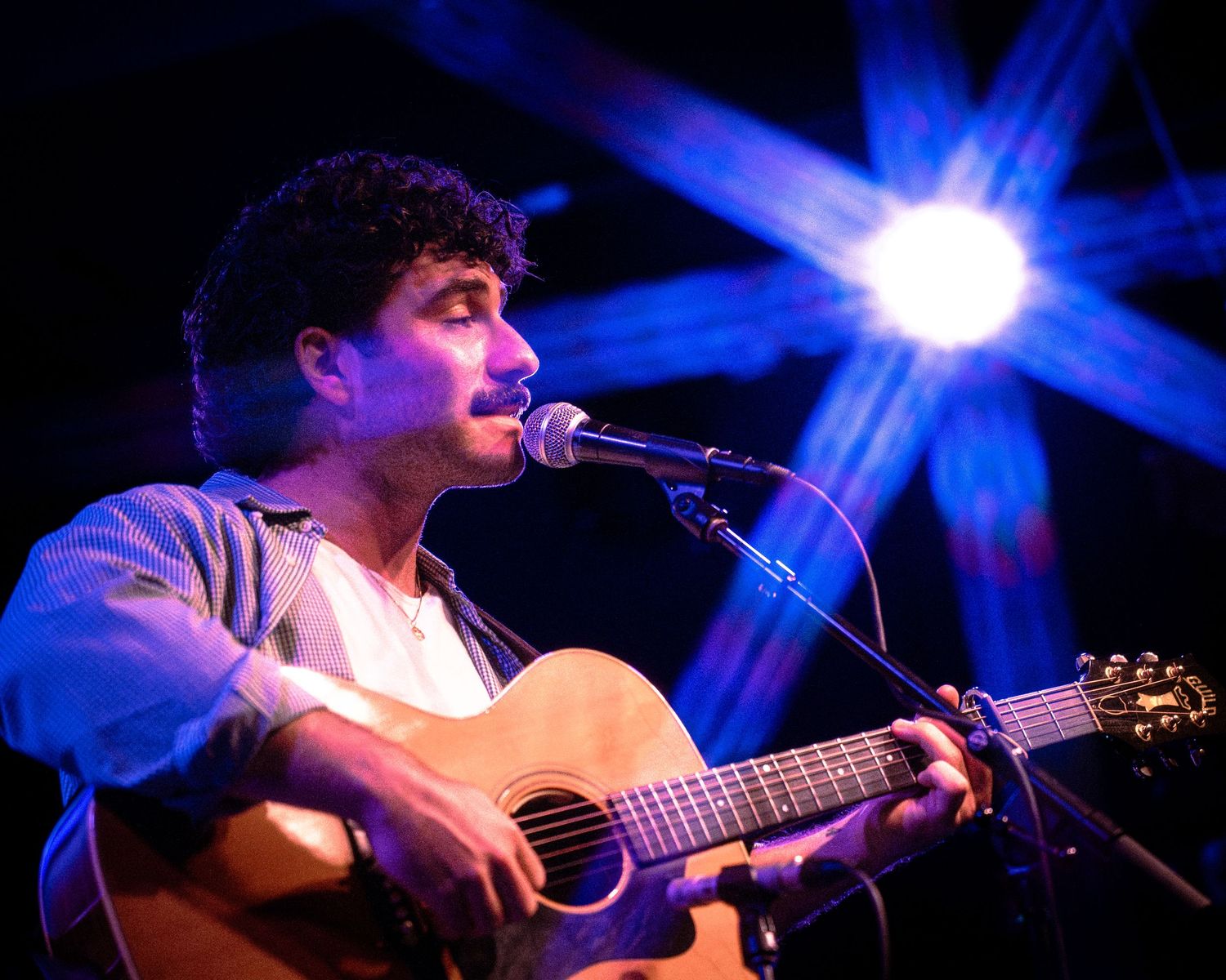 Musician performs with acoustic guitar under blue stage lighting at intimate concert venue.