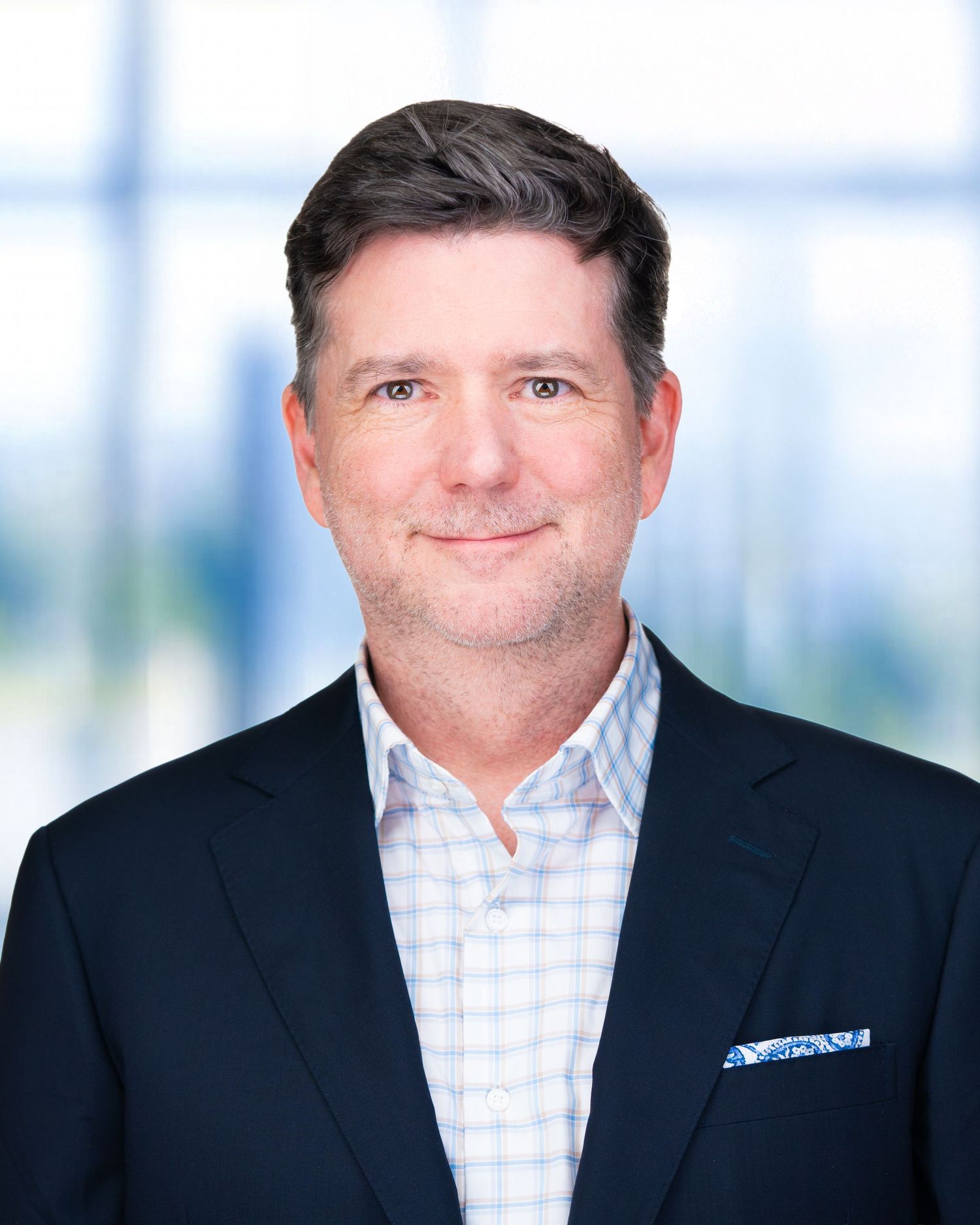 Professional portrait of person in checked shirt and dark blazer against office window background.