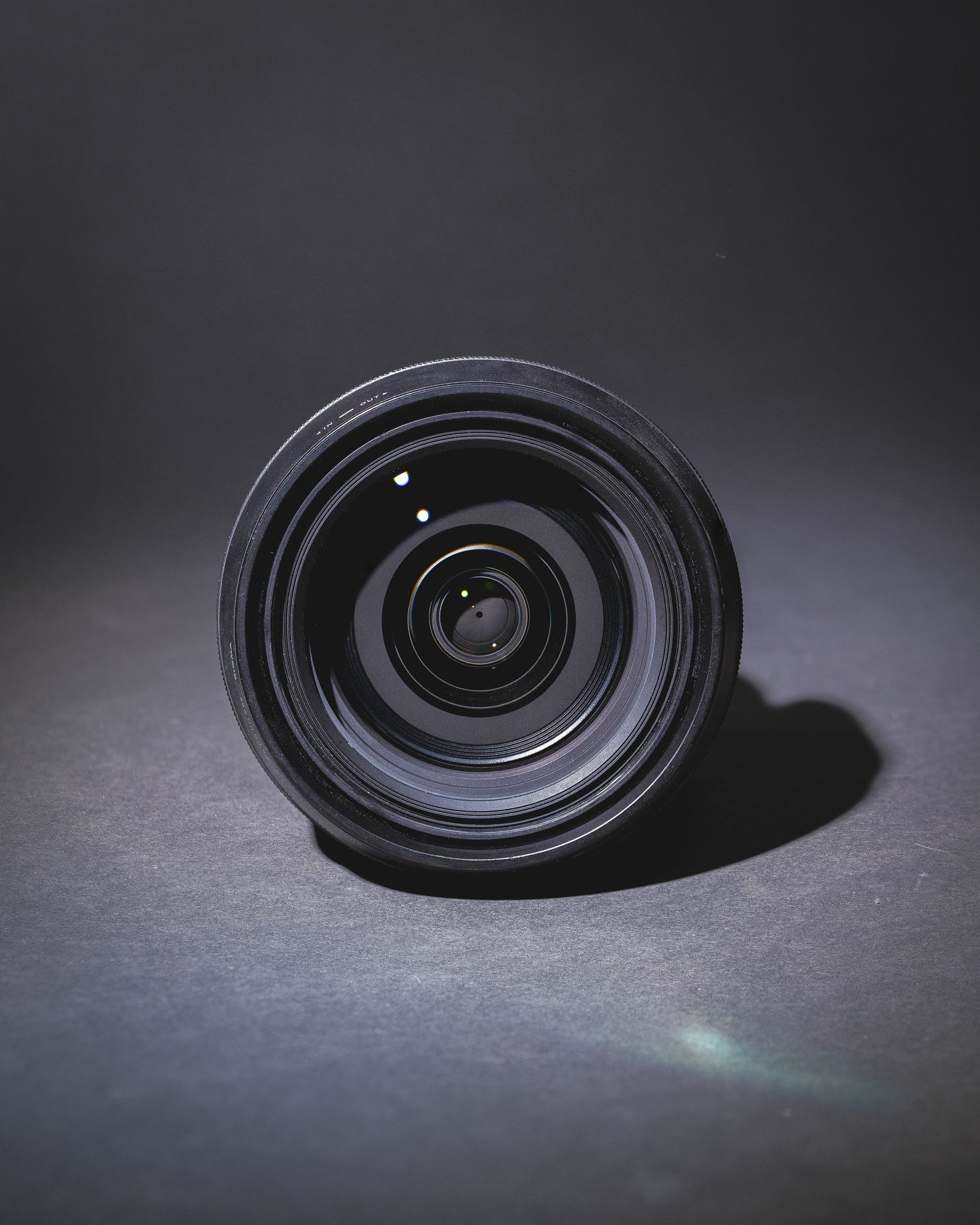 A black camera lens with glass elements sits on a dark surface with dramatic lighting and shadows.