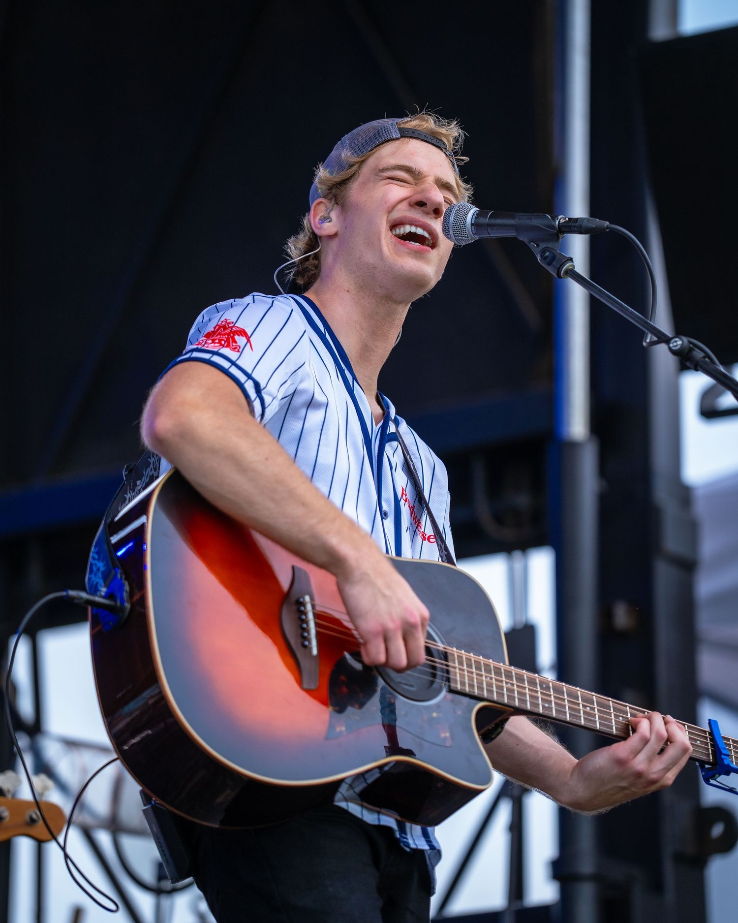Performer in pinstripe baseball jersey playing acoustic guitar on outdoor festival stage.