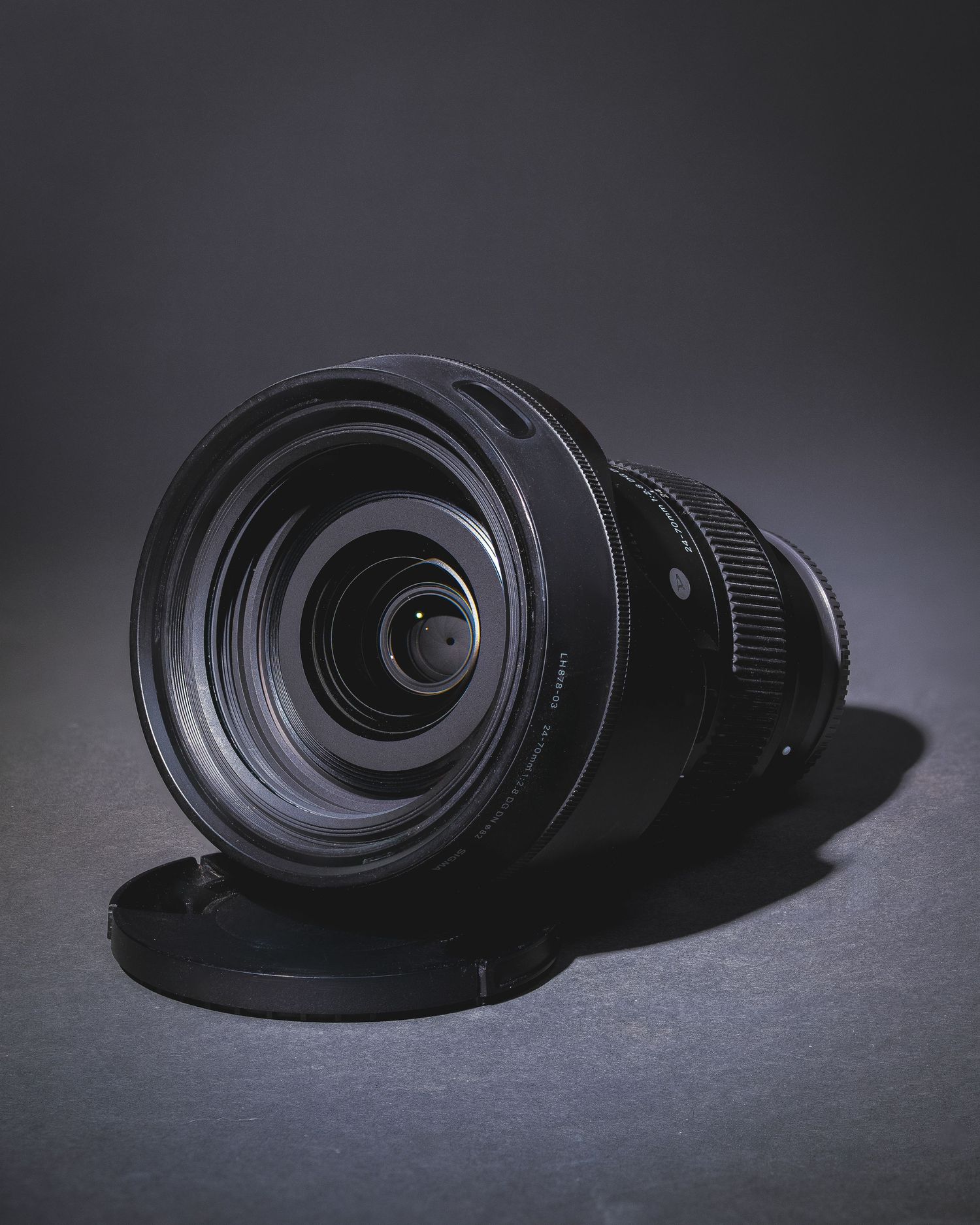A black camera lens with wide aperture sits on a dark surface against a shadowy background.