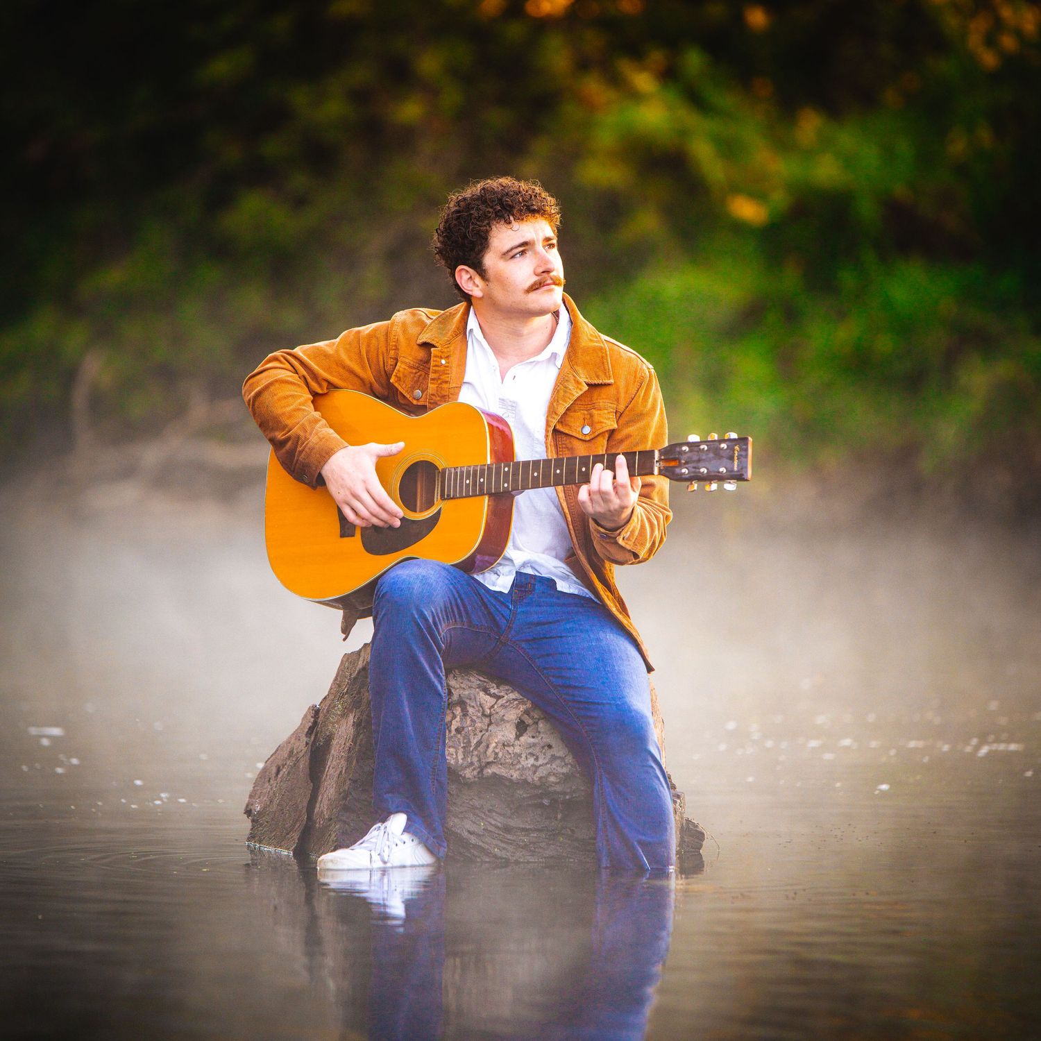 Musician in brown jacket and jeans plays acoustic guitar while seated outdoors with misty lake in background at sunset.