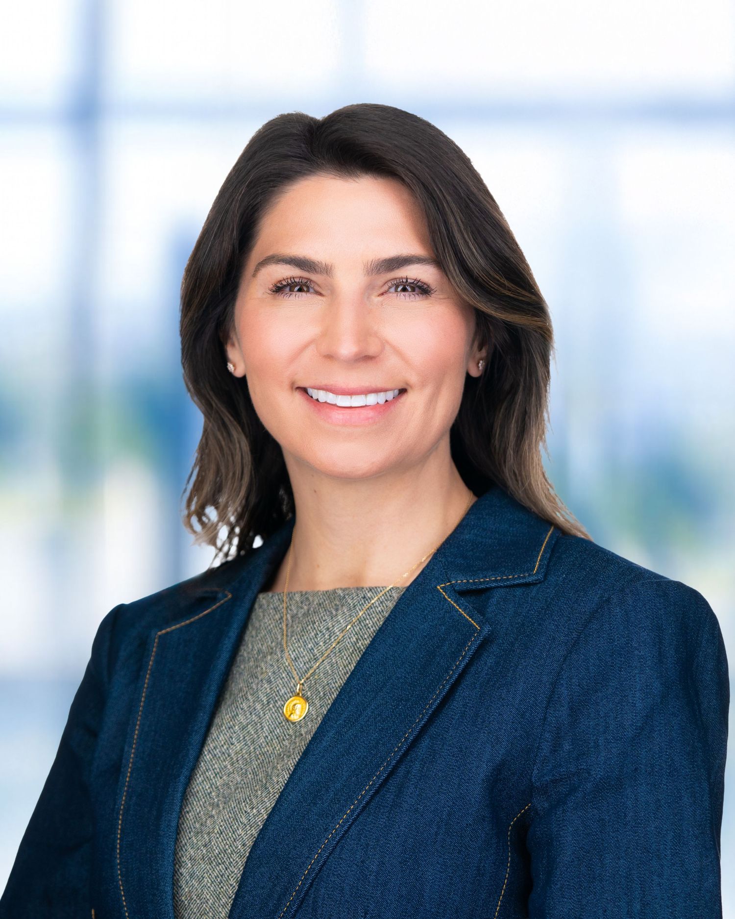Professional corporate headshot of person wearing navy blazer and olive top against bright office setting.