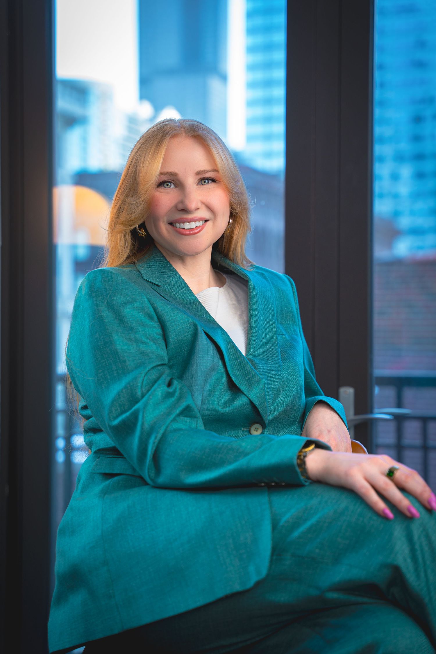 Person in vibrant turquoise blazer sitting by window with city skyline visible.
