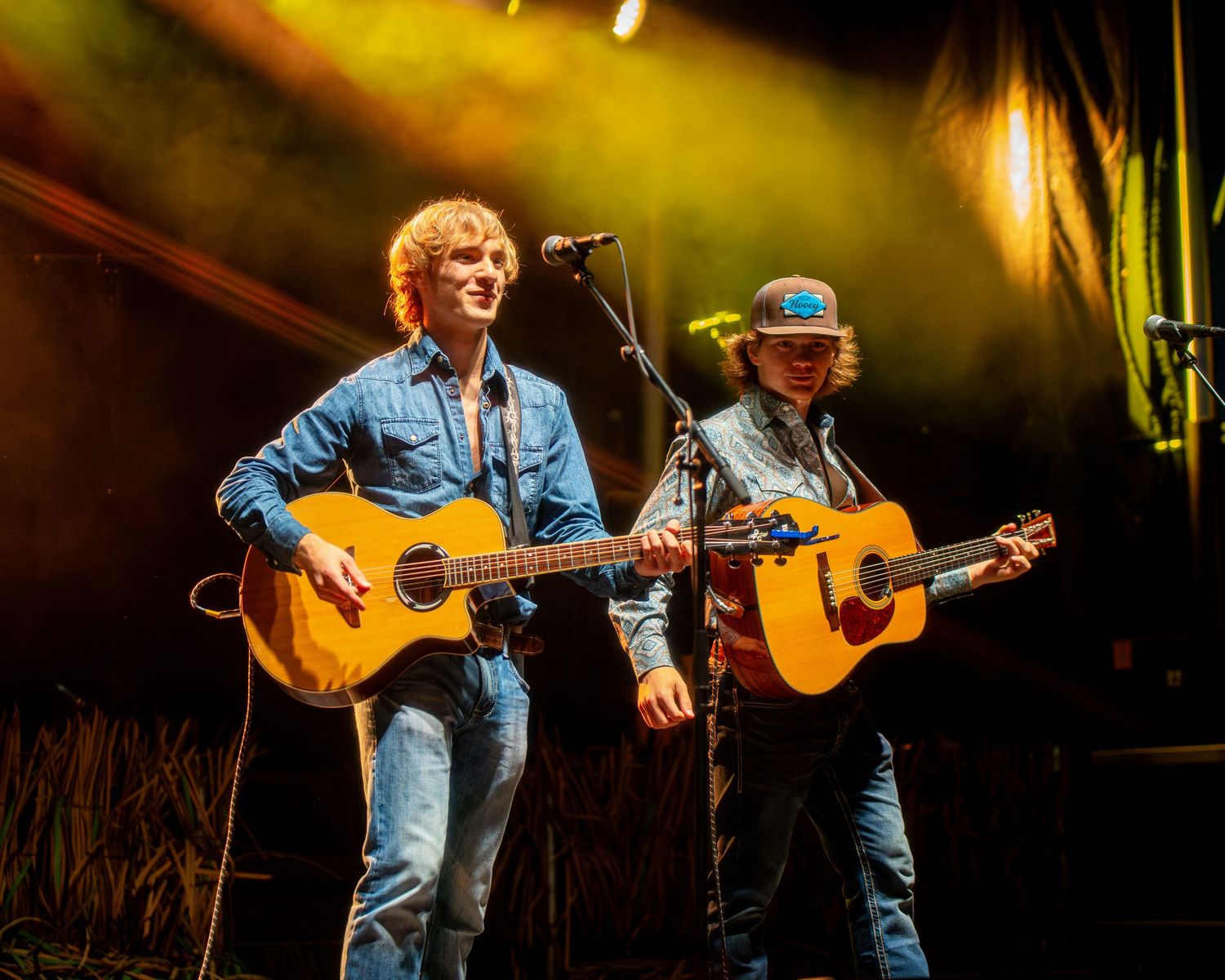 Musician performing on acoustic guitar wearing denim and overalls on stage in warm lighting.