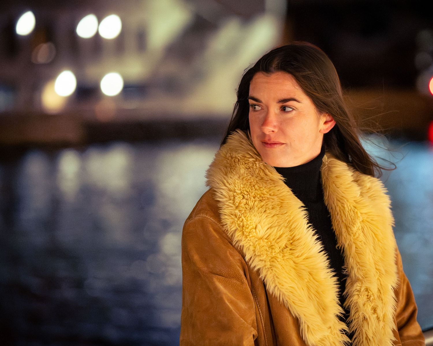 Side view portrait in a tan fur-trimmed coat against evening city lights.