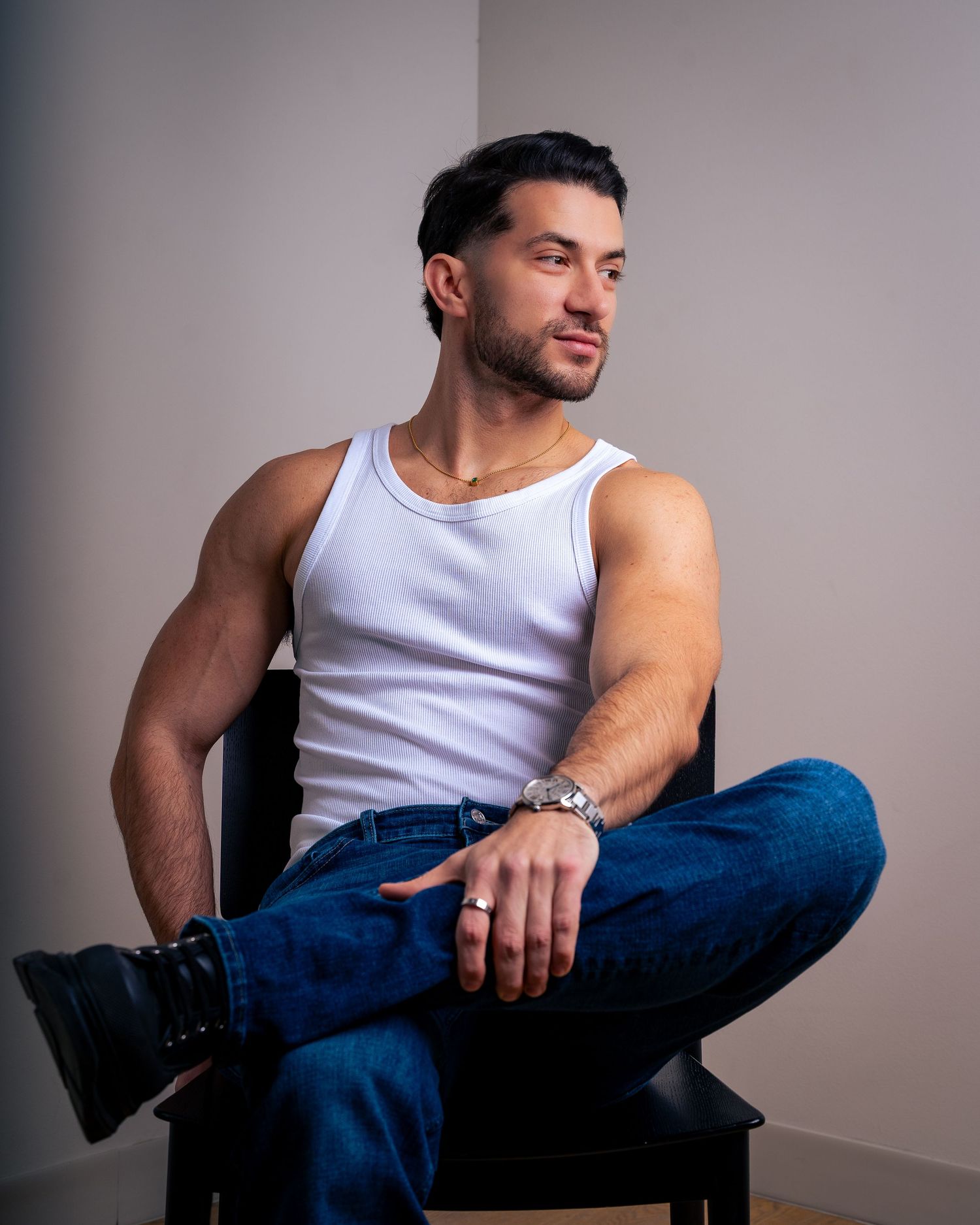 Athletic figure in white tank top and jeans poses casually on chair against gray backdrop.