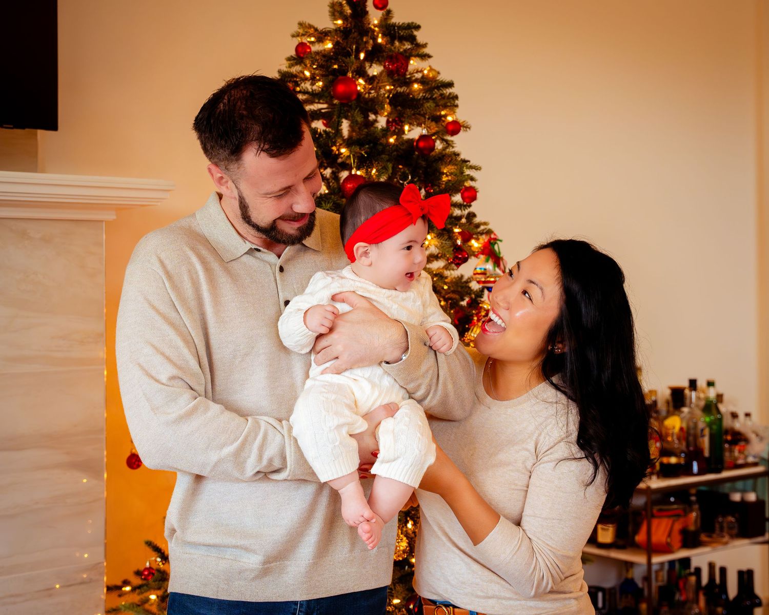 A couple shares tender moments with their newborn baby in front of a decorated Christmas tree during a holiday photoshoot.