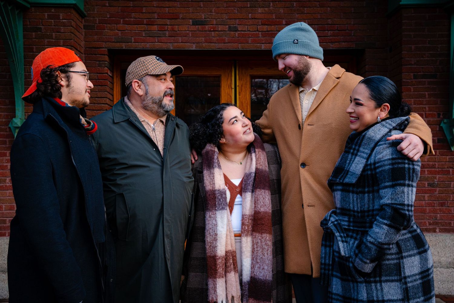 A group of five people wearing winter coats and hats stand close together against a brick wall.