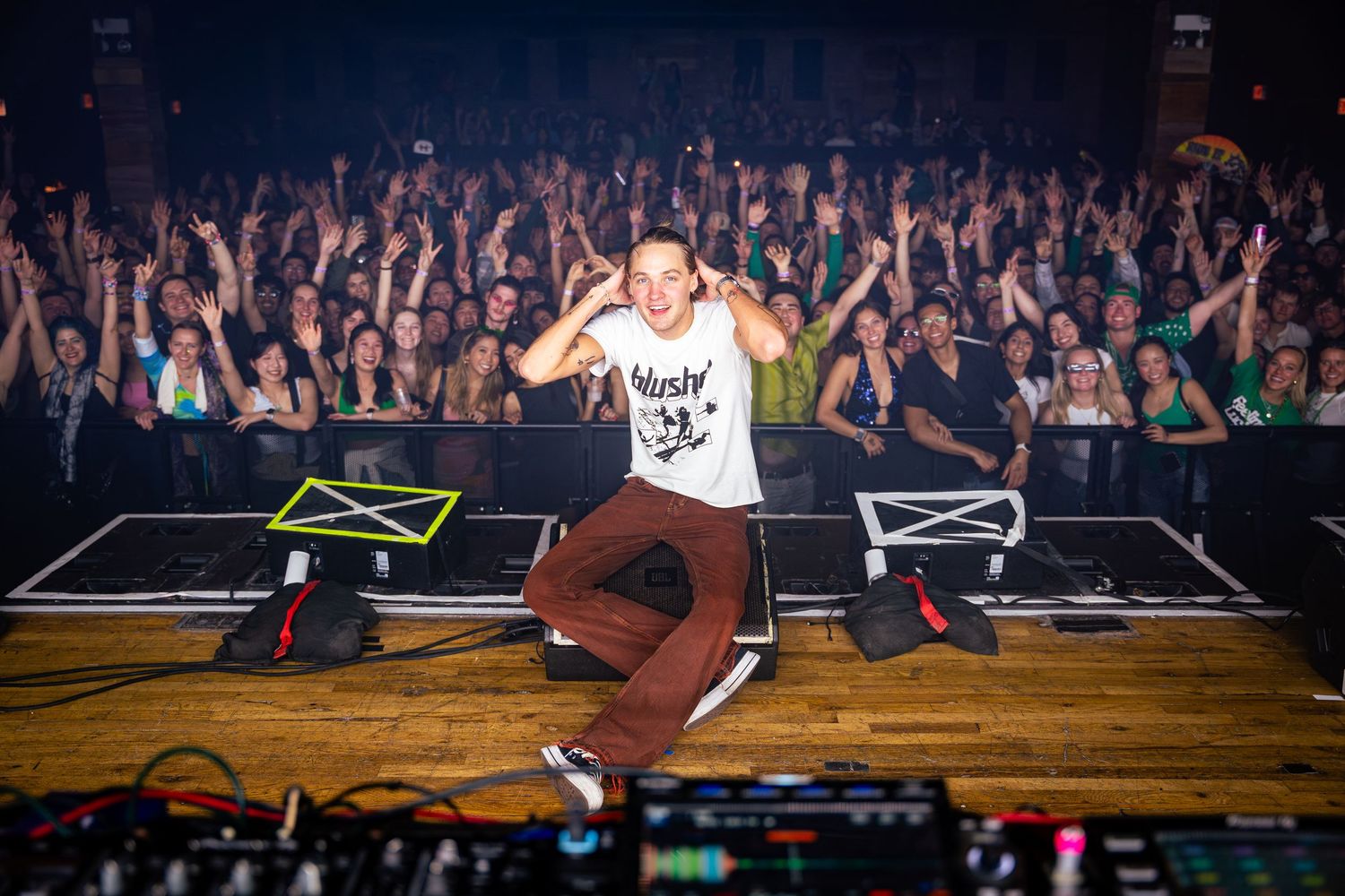 Energetic performer on stage with large crowd of fans raising hands in background.