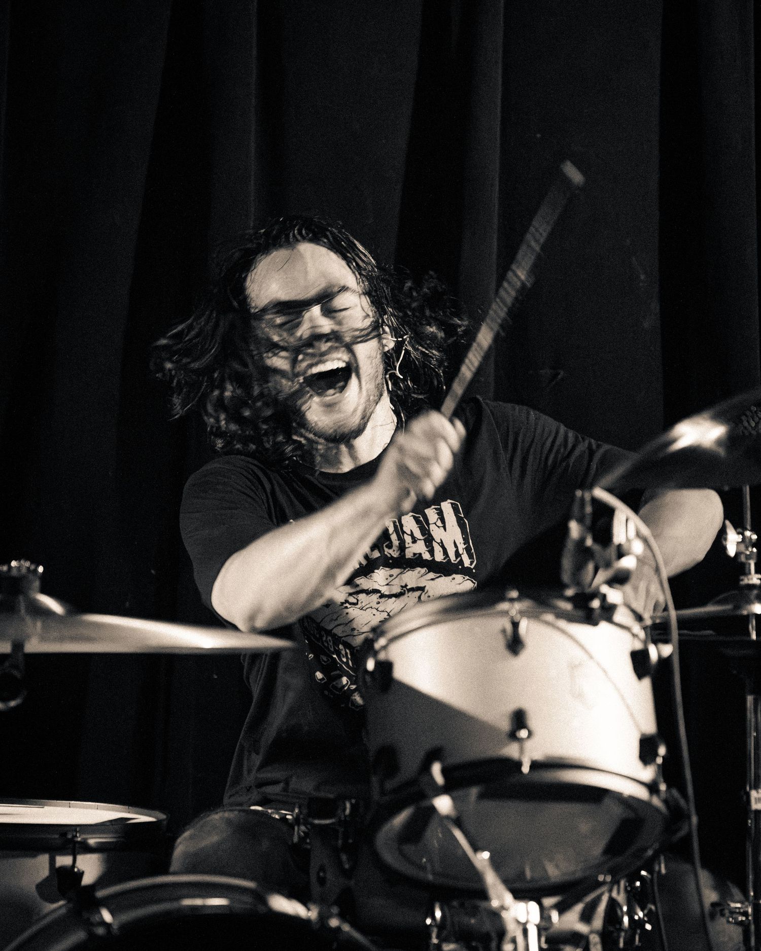 Drummer plays energetically on drum kit in dramatic black and white lighting at music venue.