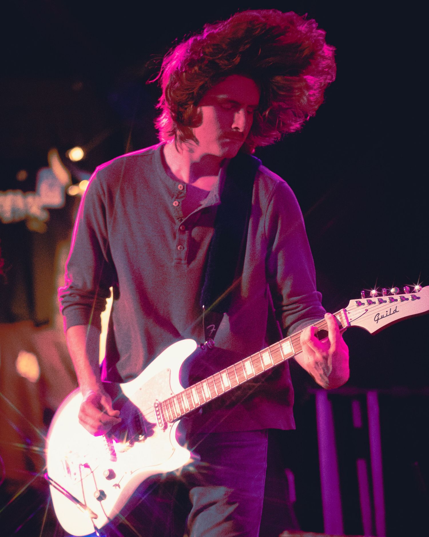 A guitarist with curly hair performs on stage under purple lighting with an electric guitar during a live concert.