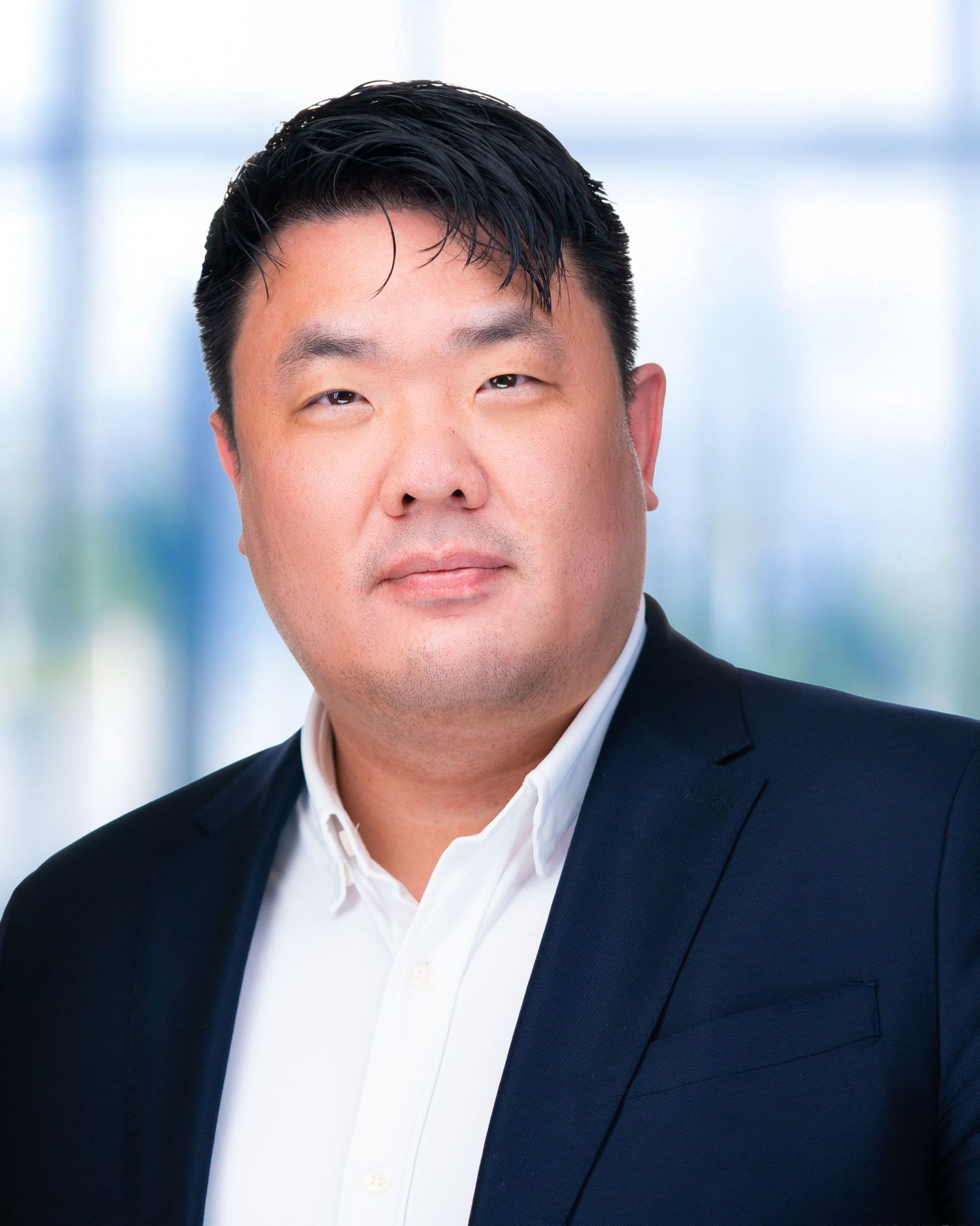 Corporate headshot of professional in dark suit and white shirt against light office background.