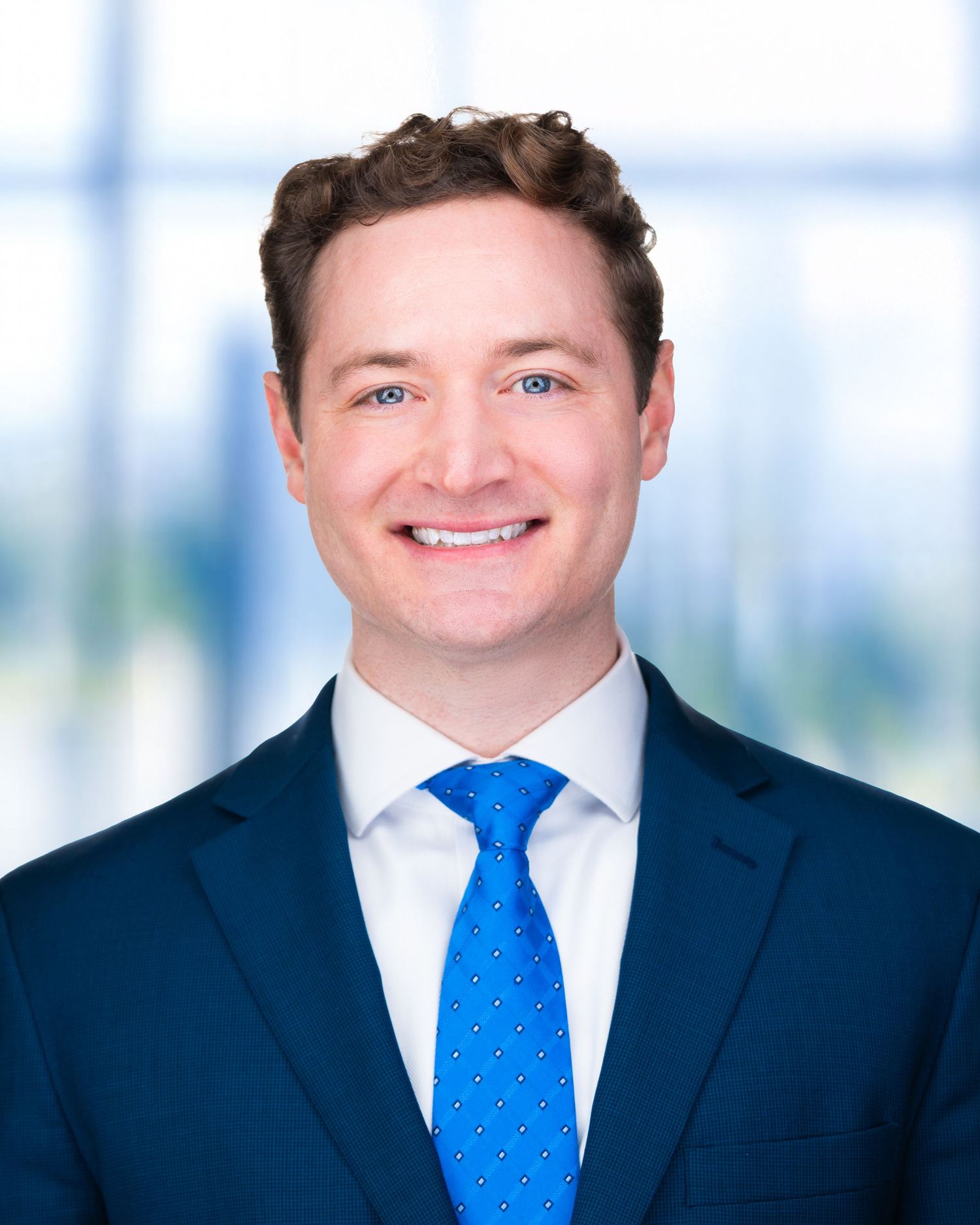 Business portrait of person wearing navy suit with bright blue tie against modern office setting.