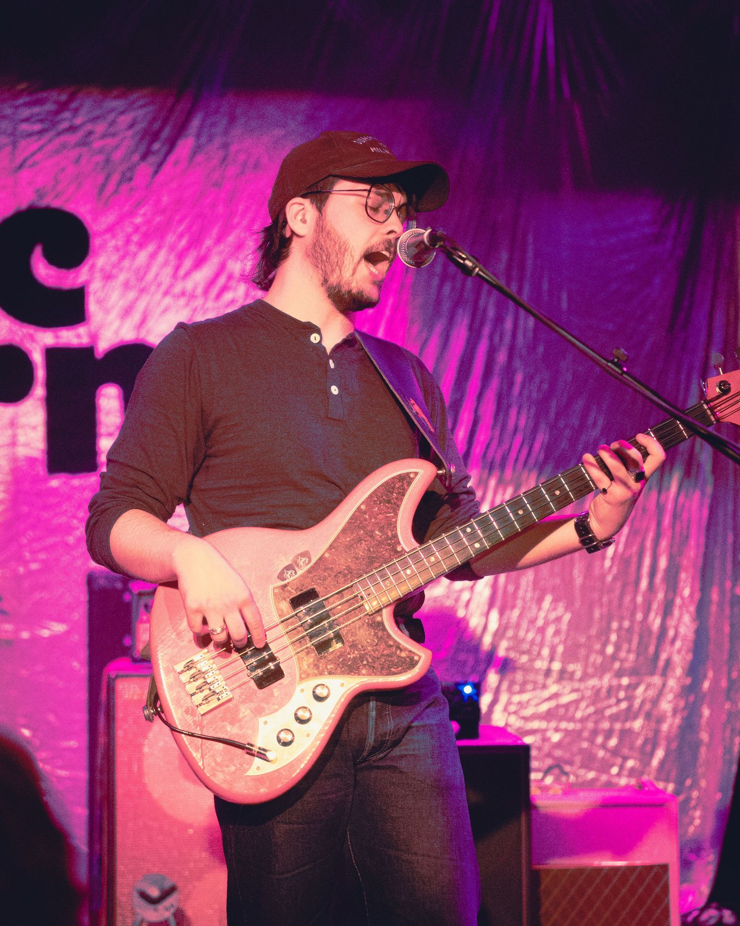 Bass guitarist performs energetically on stage with pink and purple lighting while singing into a microphone.