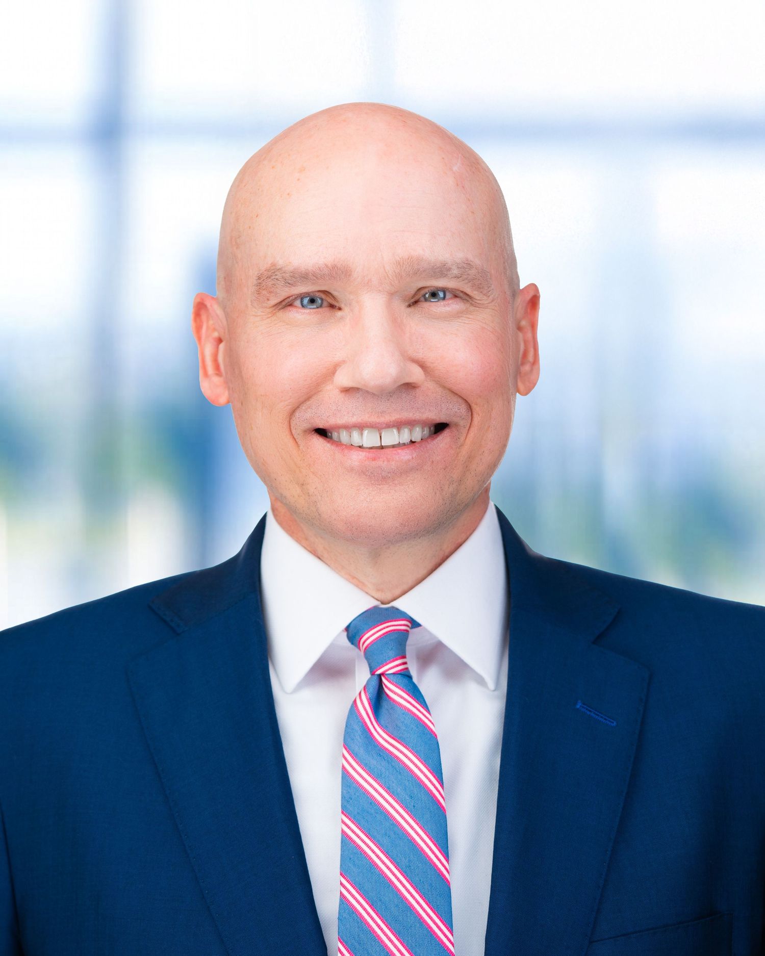 Professional headshot of person wearing striped tie and navy suit with bright background.