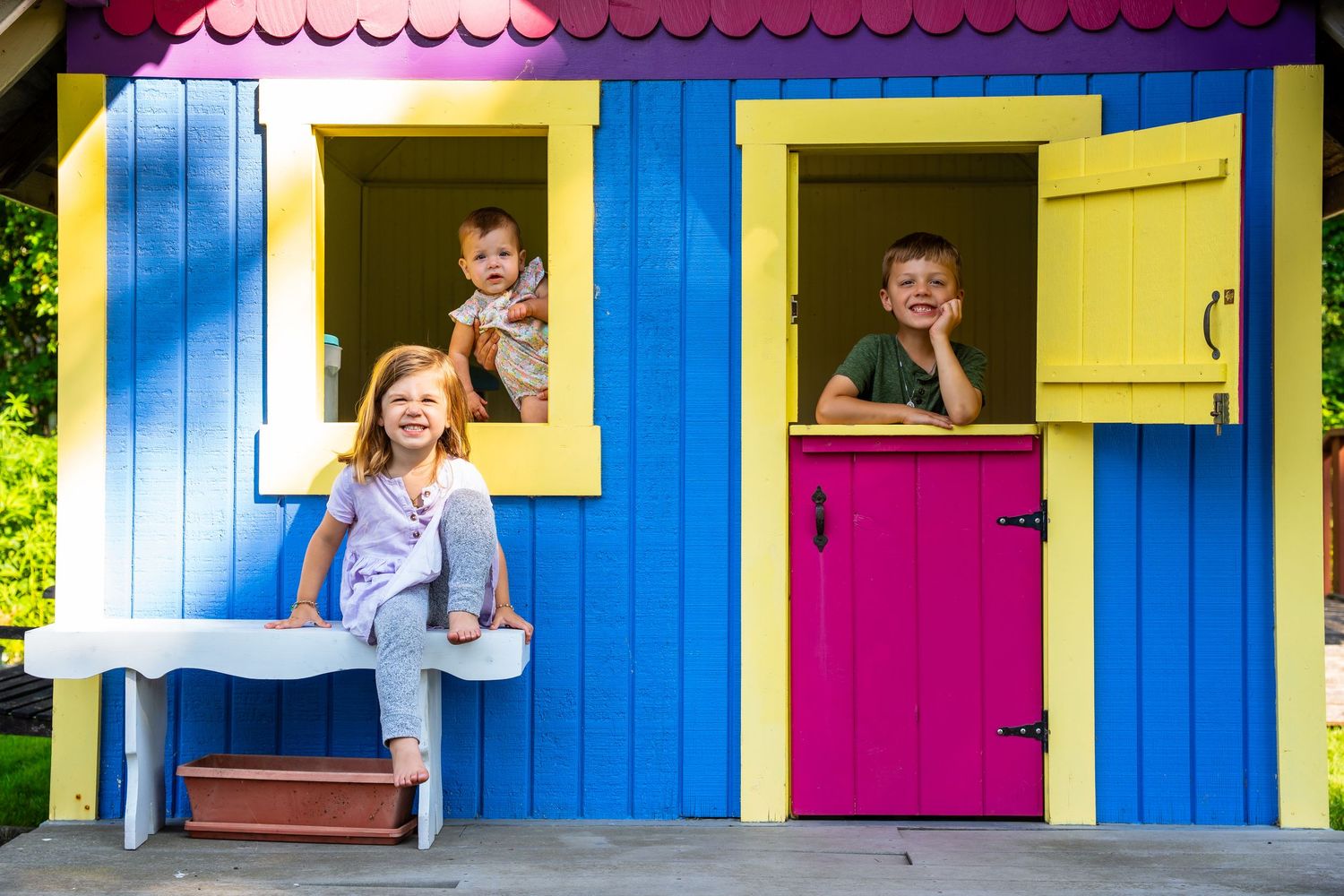 Colorful playhouse windows with pink, yellow, and blue shutters show children peeking out and smiling at the camera.