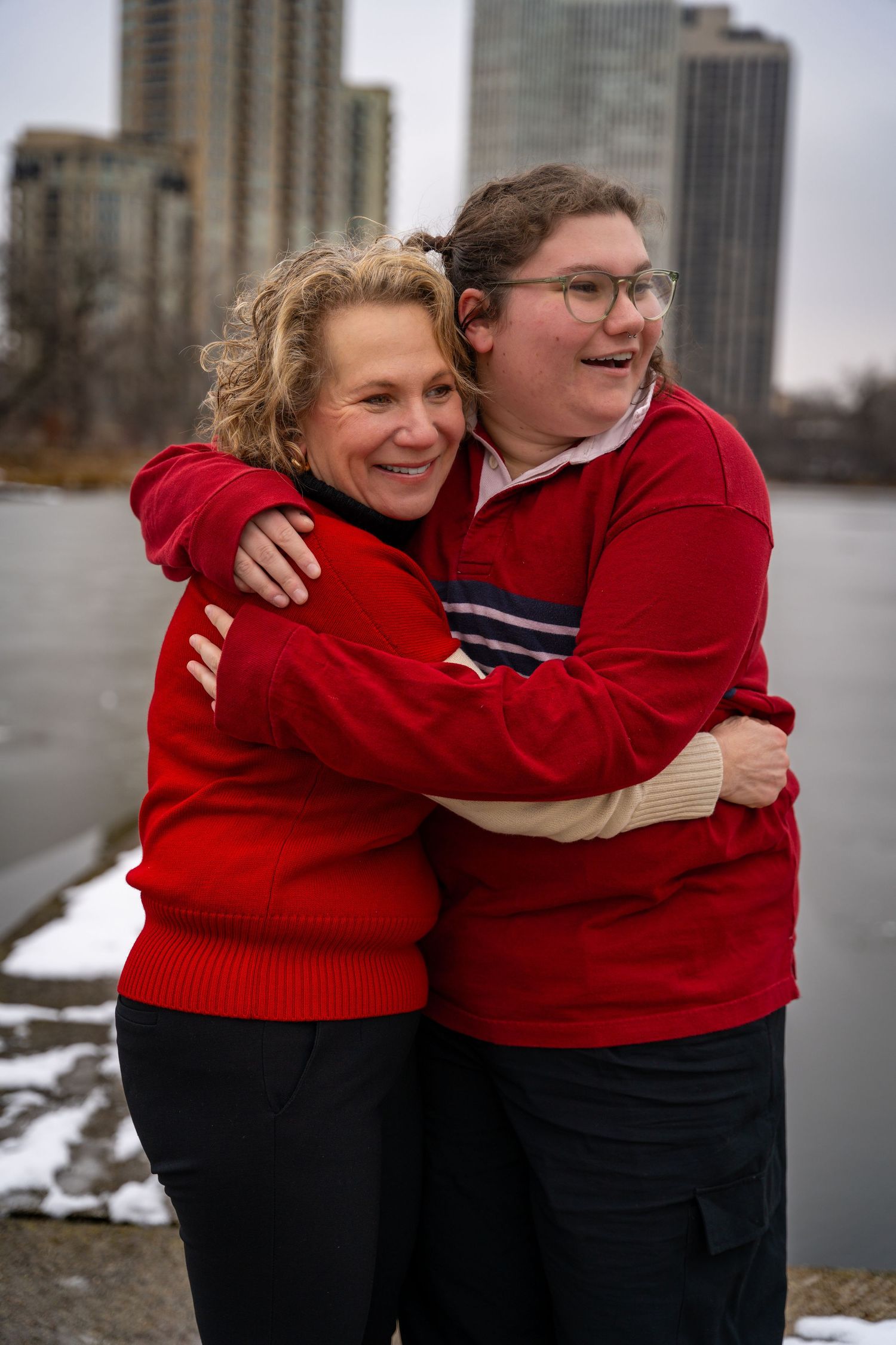 Two friends wearing matching red sweaters share a warm embrace while standing near Lake Michigan in Chicago.