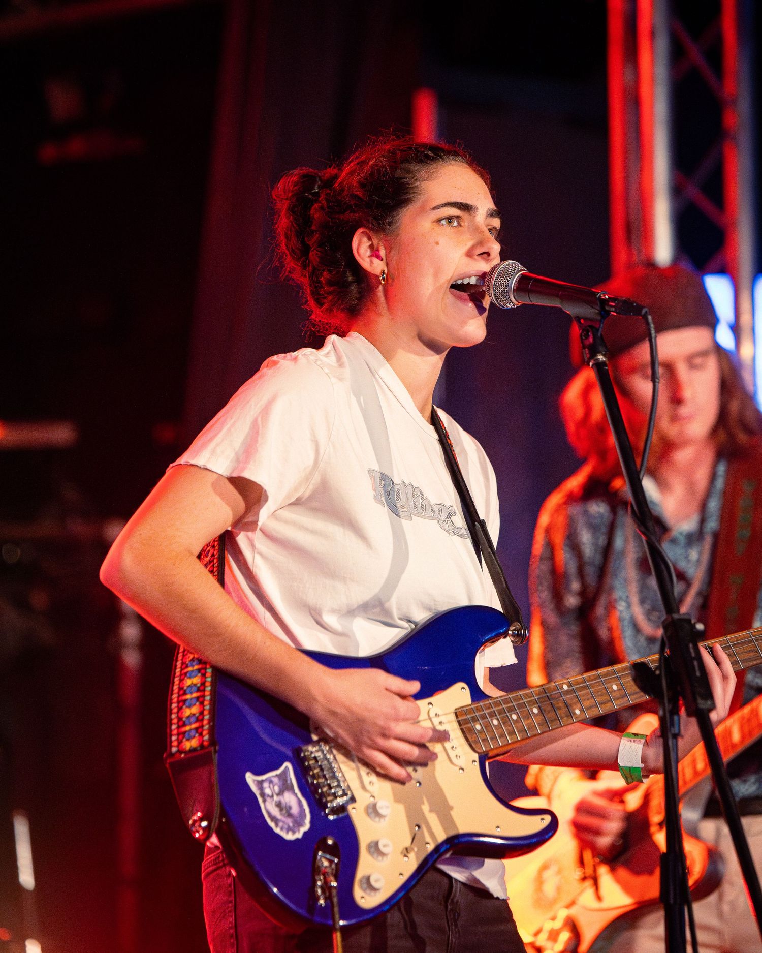 Performer strums blue electric guitar on stage illuminated by red stage lights.