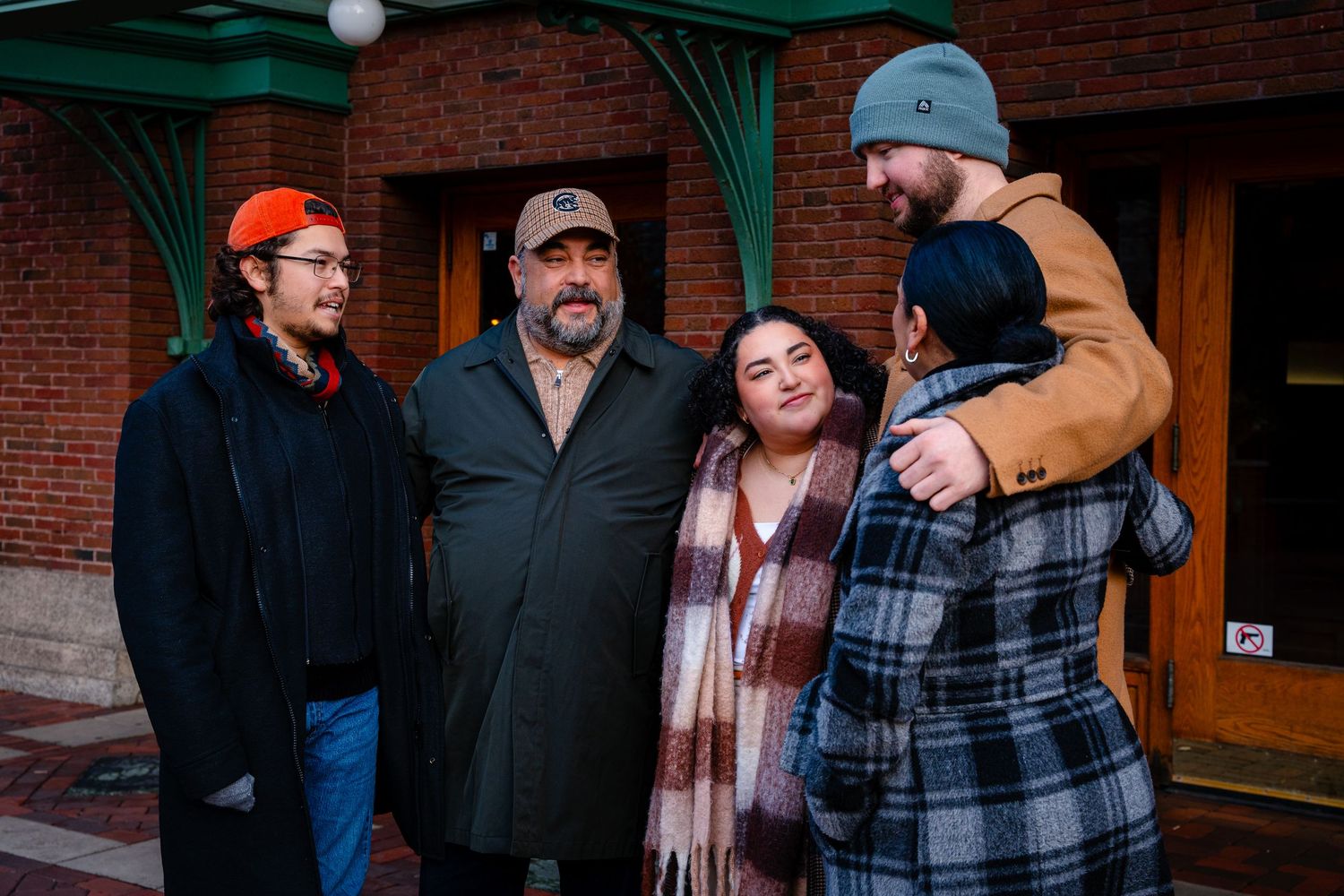 A group of people in winter clothing stand together outside a brick building with green trim.