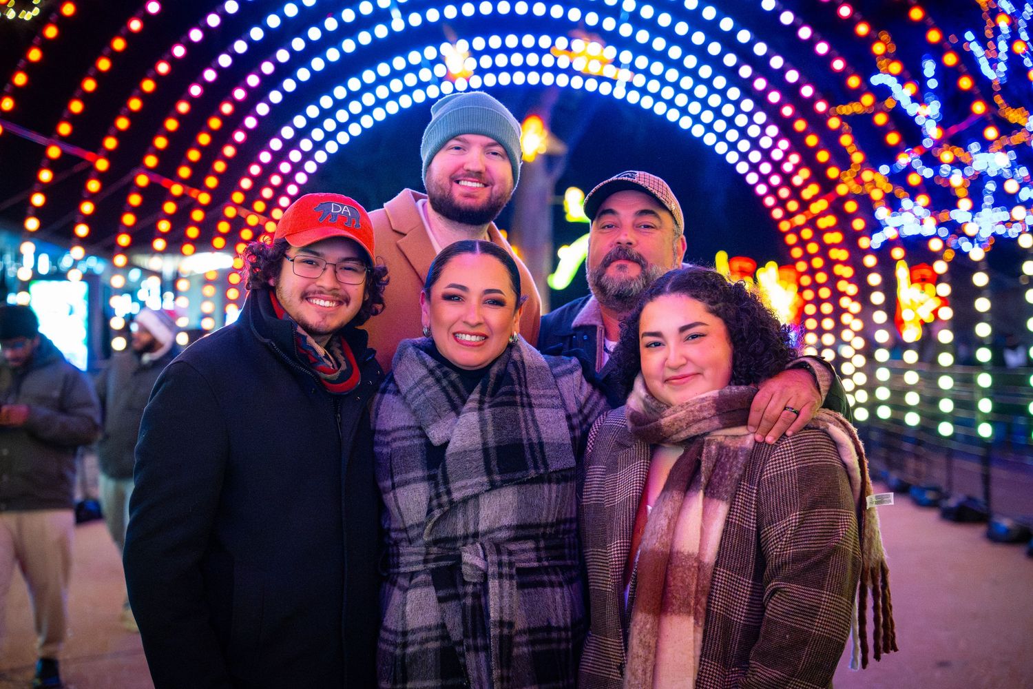 A group of friends bundled up in winter coats smile together at a holiday light display with colorful illuminated arches.