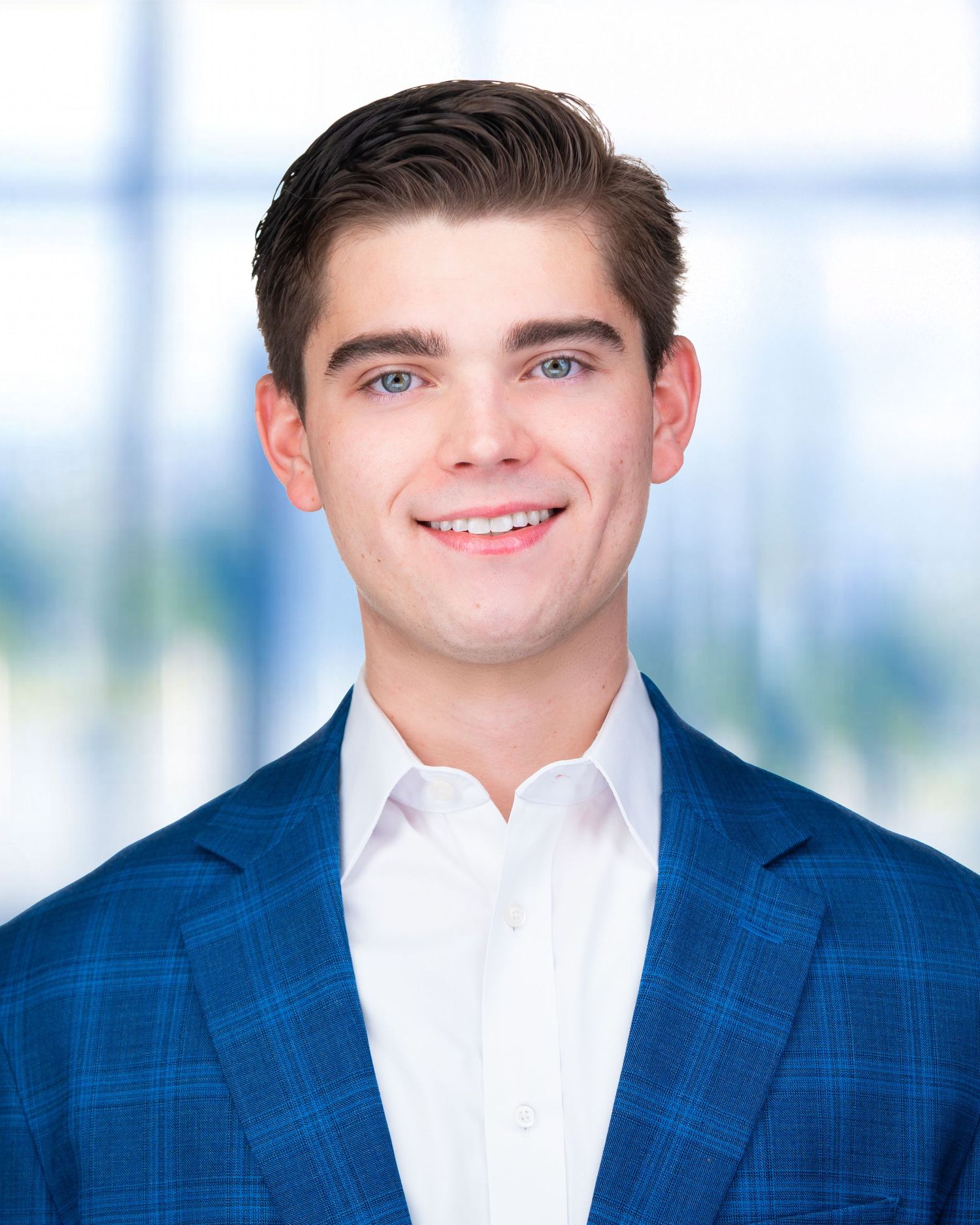 Professional headshot of person in navy blue suit jacket and white dress shirt against bright office background.