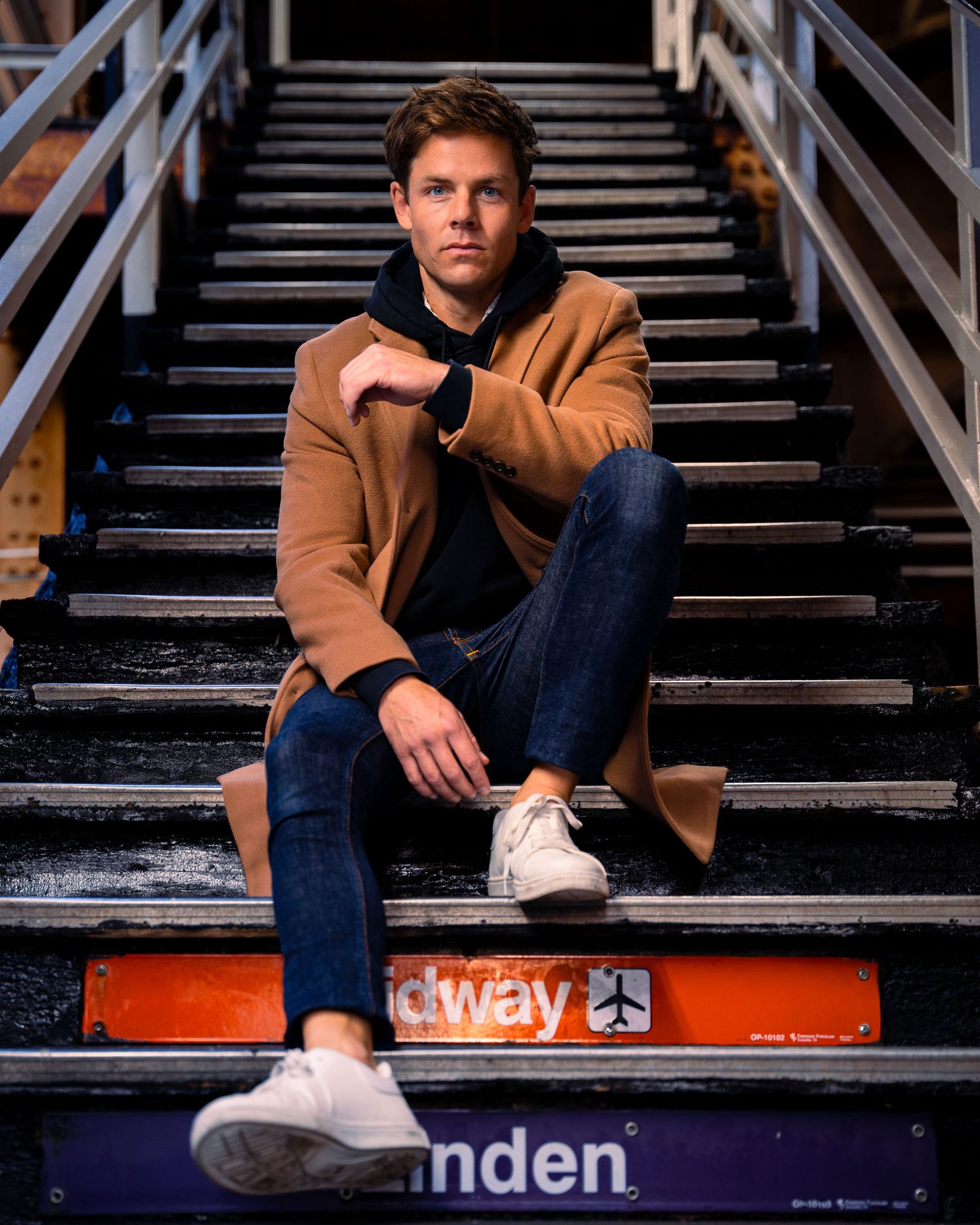 Casual menswear outfit featuring tan coat and white sneakers while sitting on urban steps.
