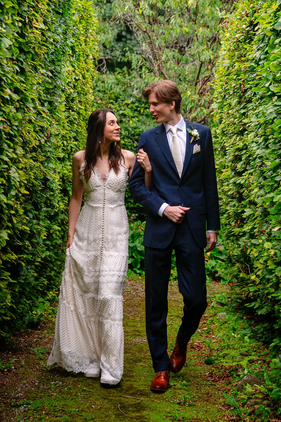 Couple in wedding attire walking through lush green garden pathway.