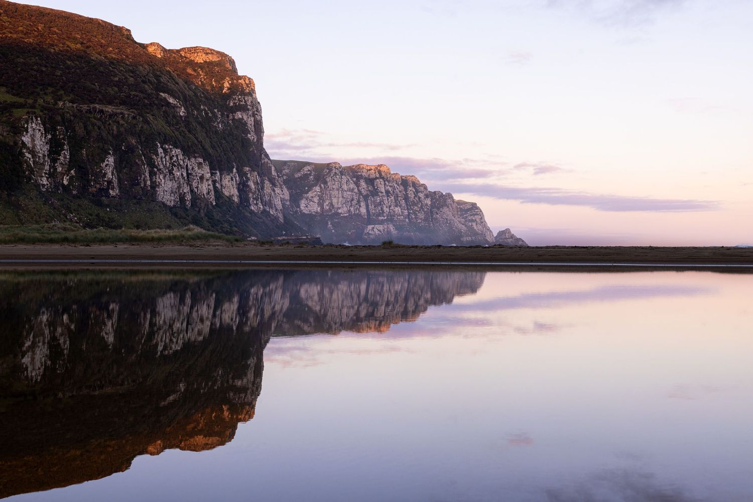 Dramatic coastal cliffs and mountains reflect in calm ocean waters at sunset creating a stunning natural panorama.