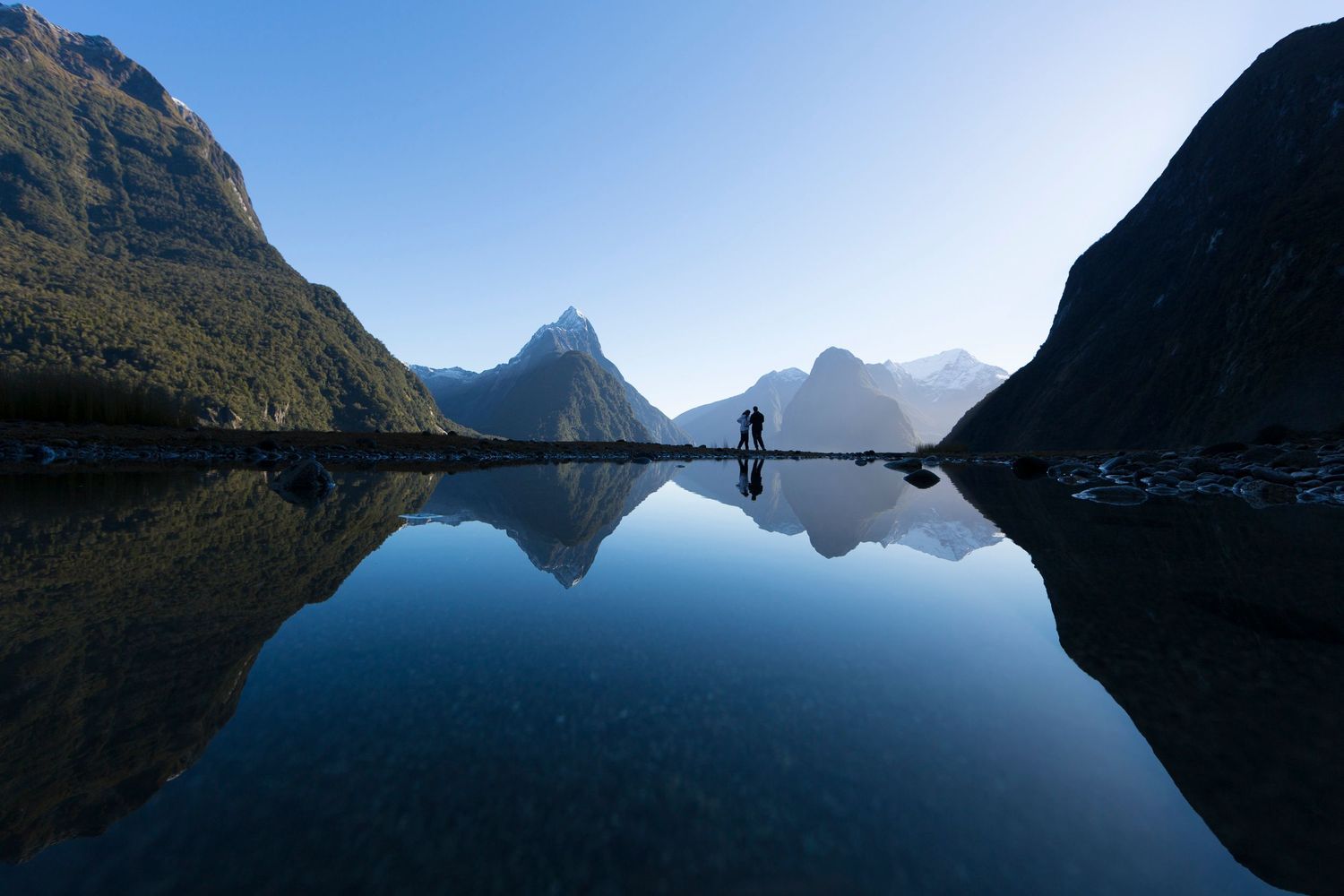 Silhouette of hiker standing on rocks at Milford Sound with mountains reflecting in still waters.
