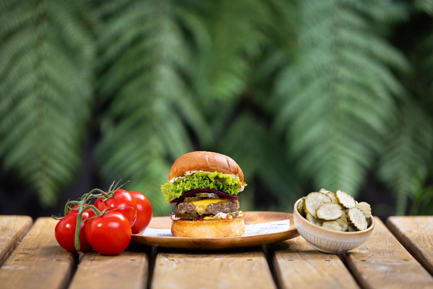 A juicy hamburger on a wooden board with fresh tomatoes and pickles against fern leaves.
